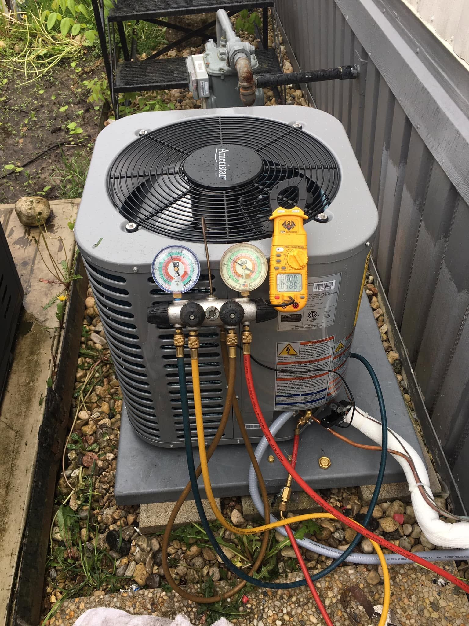 Outdoor HVAC condenser unit with gauges and hoses attached, on a gravel side yard.