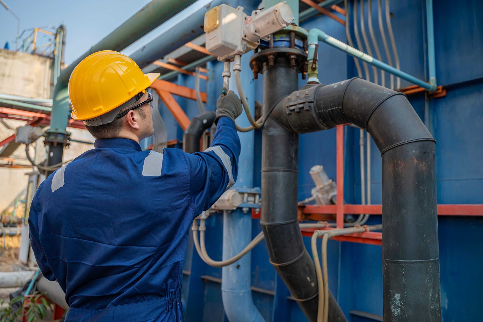 Un ouvrier portant un casque jaune et une combinaison bleue inspecte des canalisations industrielles sur un site extérieur.