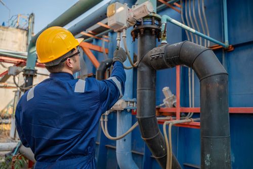 Une personne portant un casque jaune et une combinaison bleue inspecte des tuyaux sur un équipement industriel.