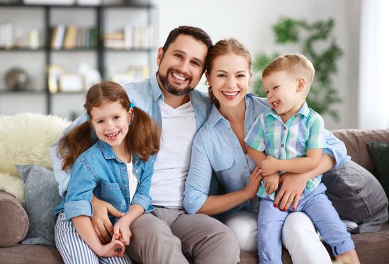 Une famille de quatre personnes sourit ensemble sur un canapé dans le salon ; deux enfants enlacent leurs parents.