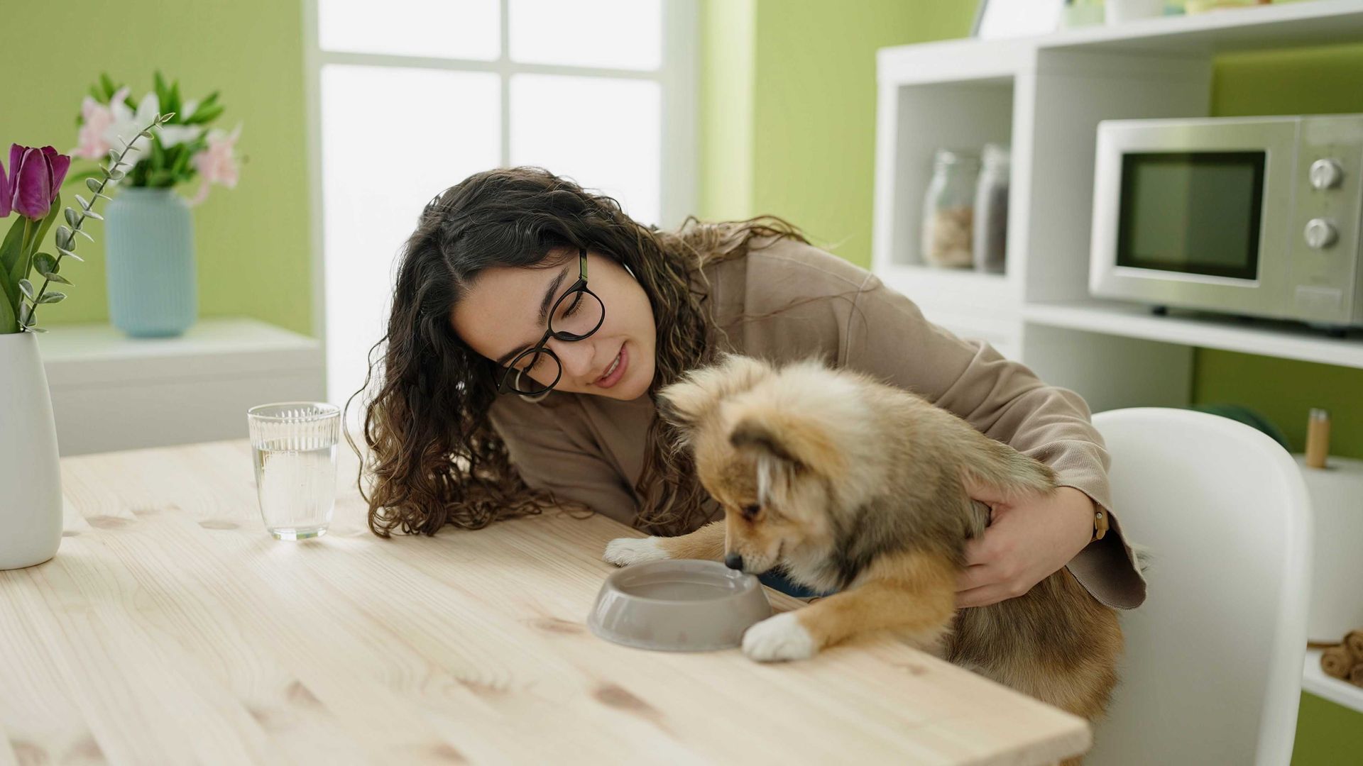 Une femme donne à manger à un chien assis à une table en bois. Le chien mange dans une gamelle tandis que la femme le caresse.