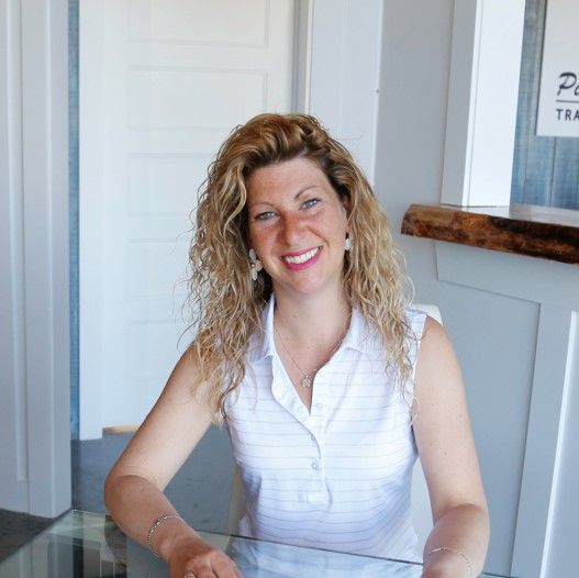 Une femme aux cheveux blonds bouclés sourit, vêtue d'un débardeur blanc. Elle est assise à l'intérieur, près d'une table en verre.