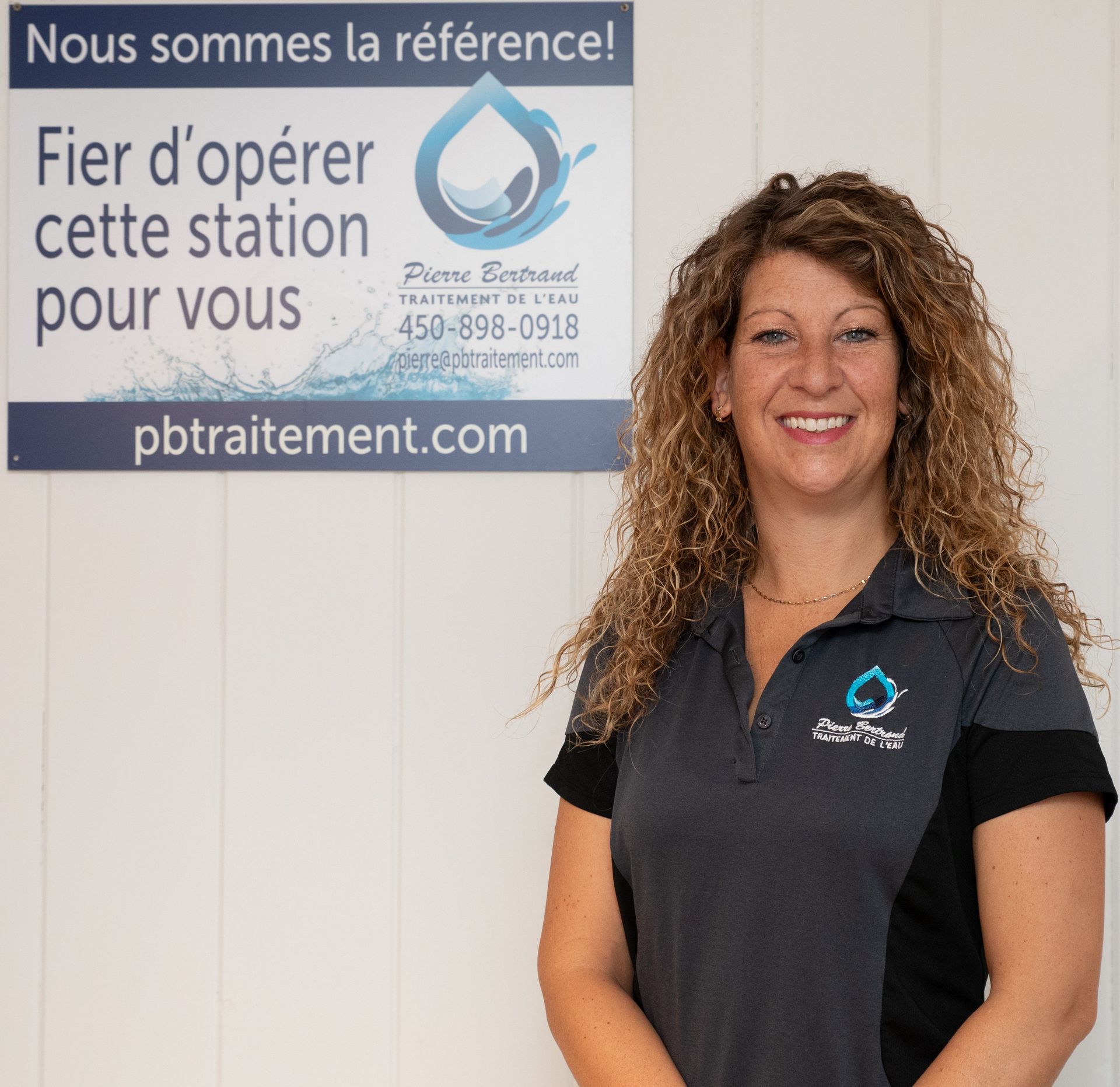 Femme souriante, debout près d'un panneau portant le logo d'une entreprise de traitement des eaux, vêtue d'une chemise grise, cheveux clairs.