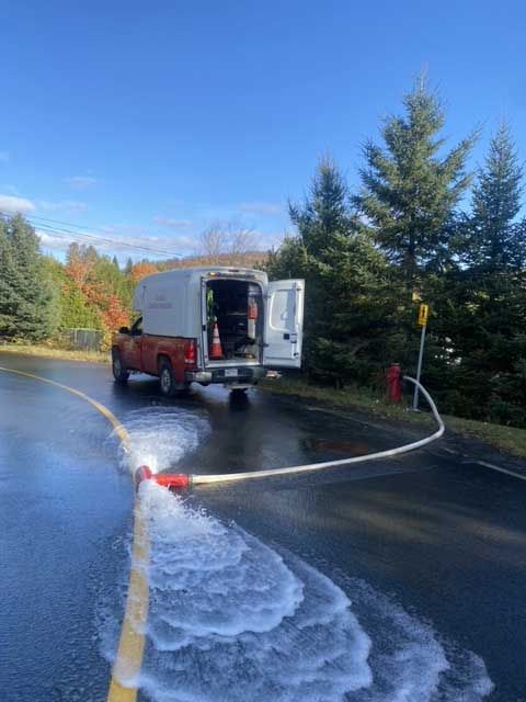 Une camionnette utilitaire rouge, portes ouvertes, avec un tuyau d'arrosage pulvérisant de la mousse sur une route mouillée.