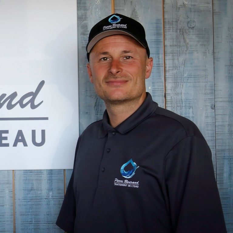 Un homme en casquette et polo se tient près d'un panneau. Il sourit. Fond : lambris bleu.