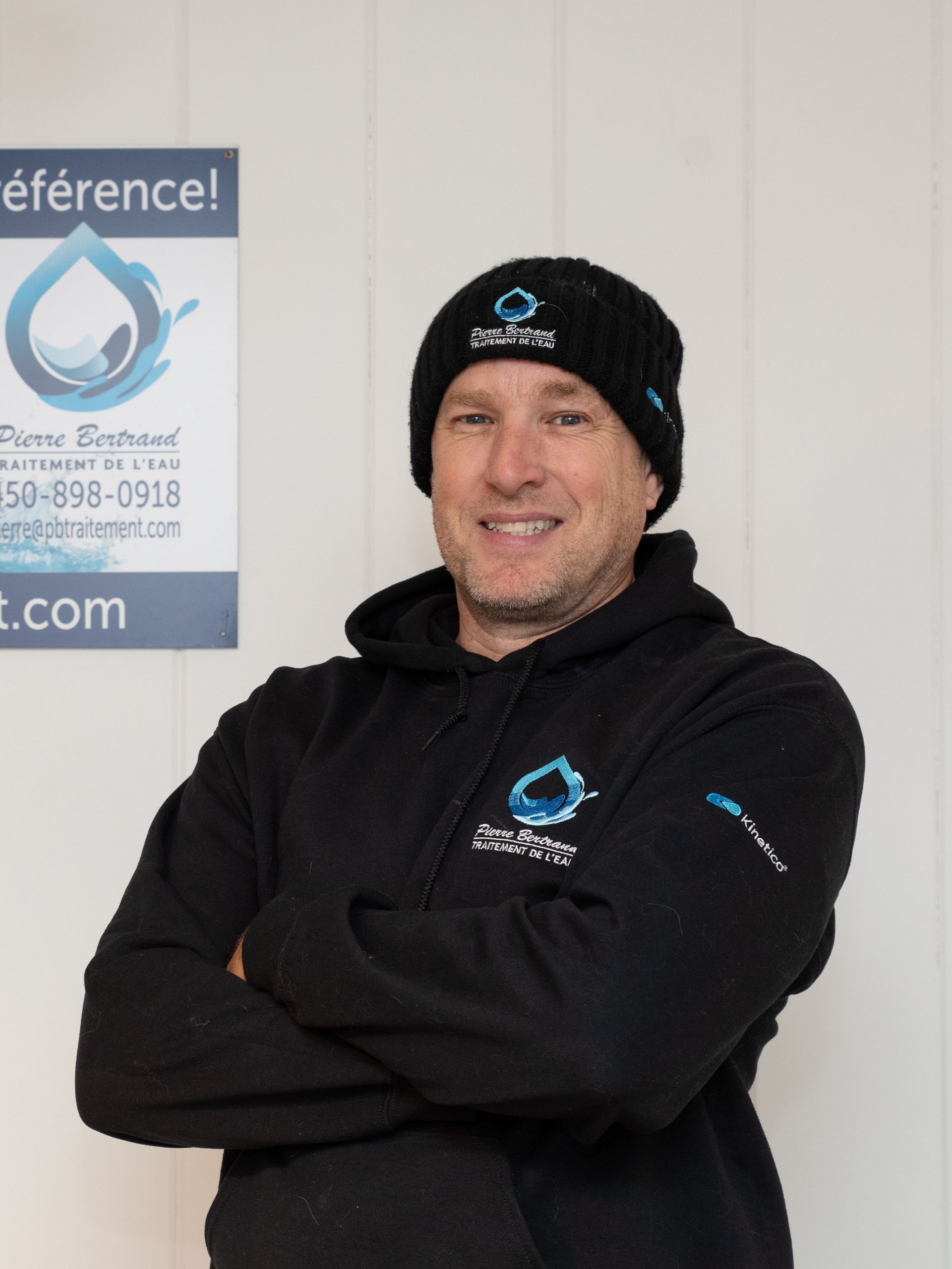 Homme en sweat à capuche et bonnet noirs, bras croisés, souriant. Panneau avec logo en forme de goutte d'eau en arrière-plan.