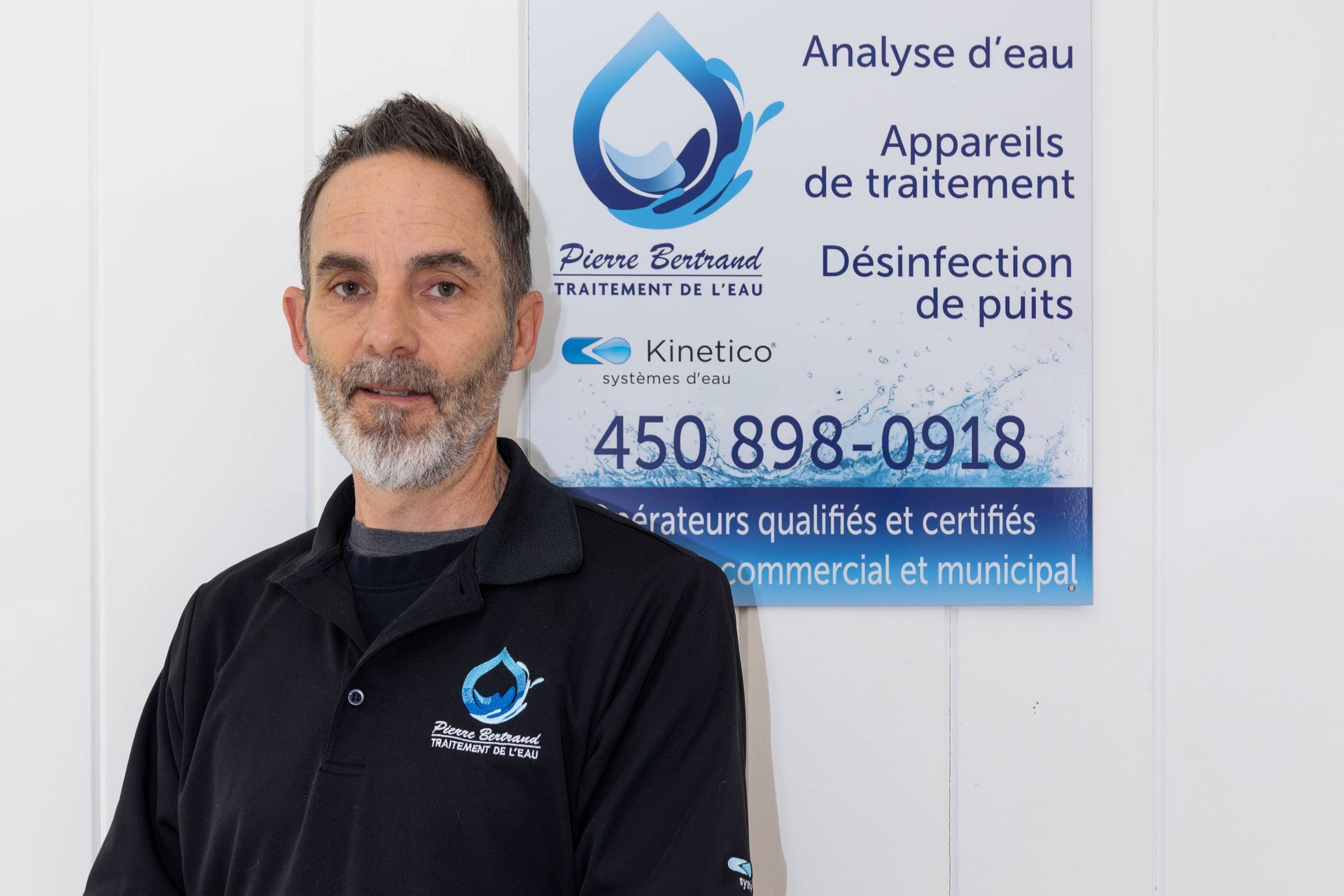 Un homme en polo noir se tient à côté d'un panneau indiquant des services liés à l'eau et un numéro de téléphone.
