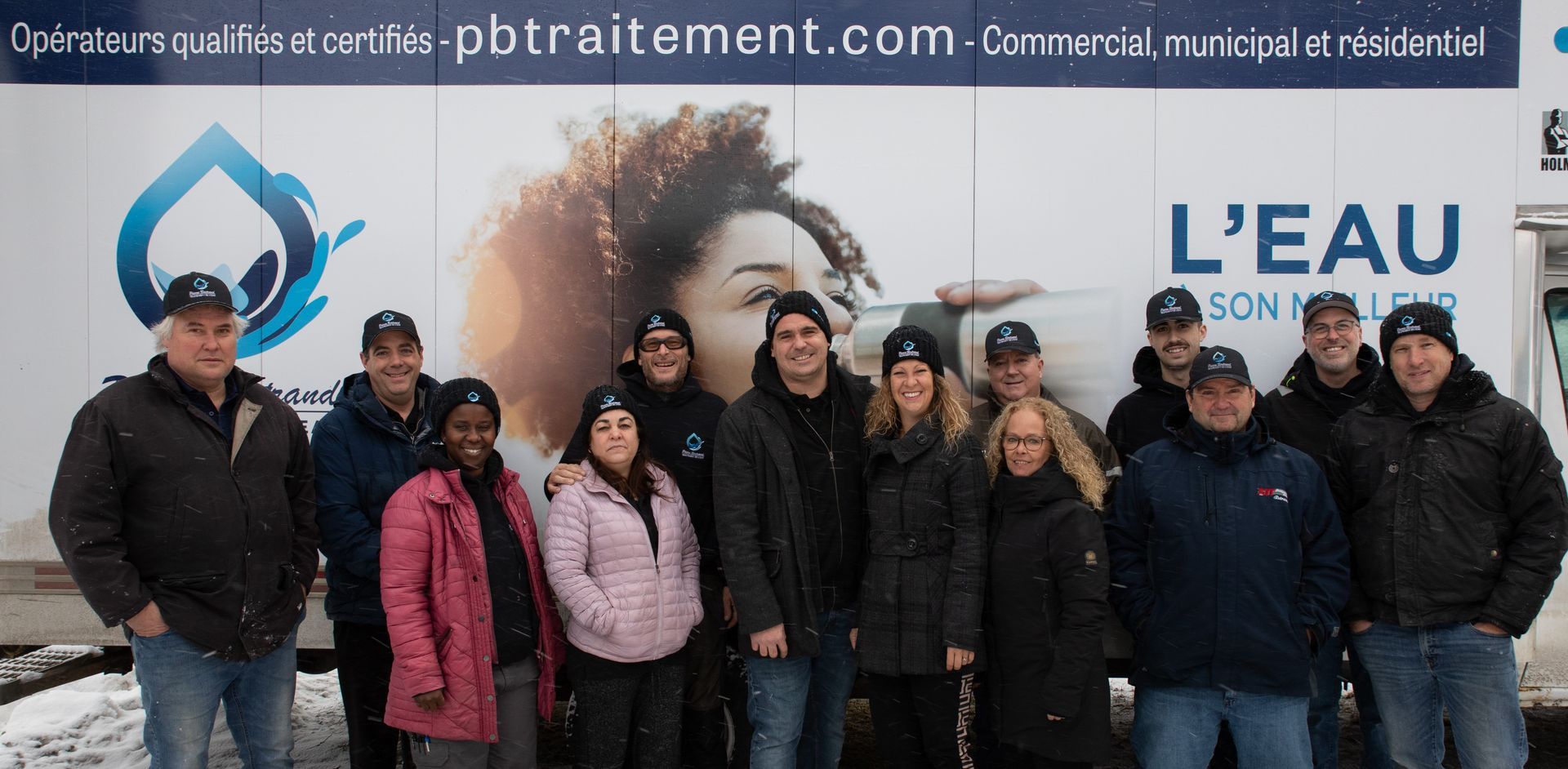 Un groupe de personnes pose devant le logo d'une entreprise de traitement des eaux. Elles portent des chapeaux et se tiennent devant un mur où est inscrit « L'EAU ».