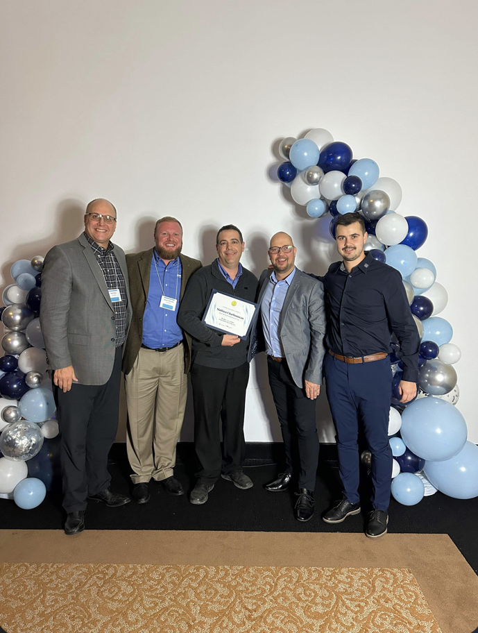 Cinq hommes, debout, posent près d'une arche de ballons, tenant un prix. Ils sourient.