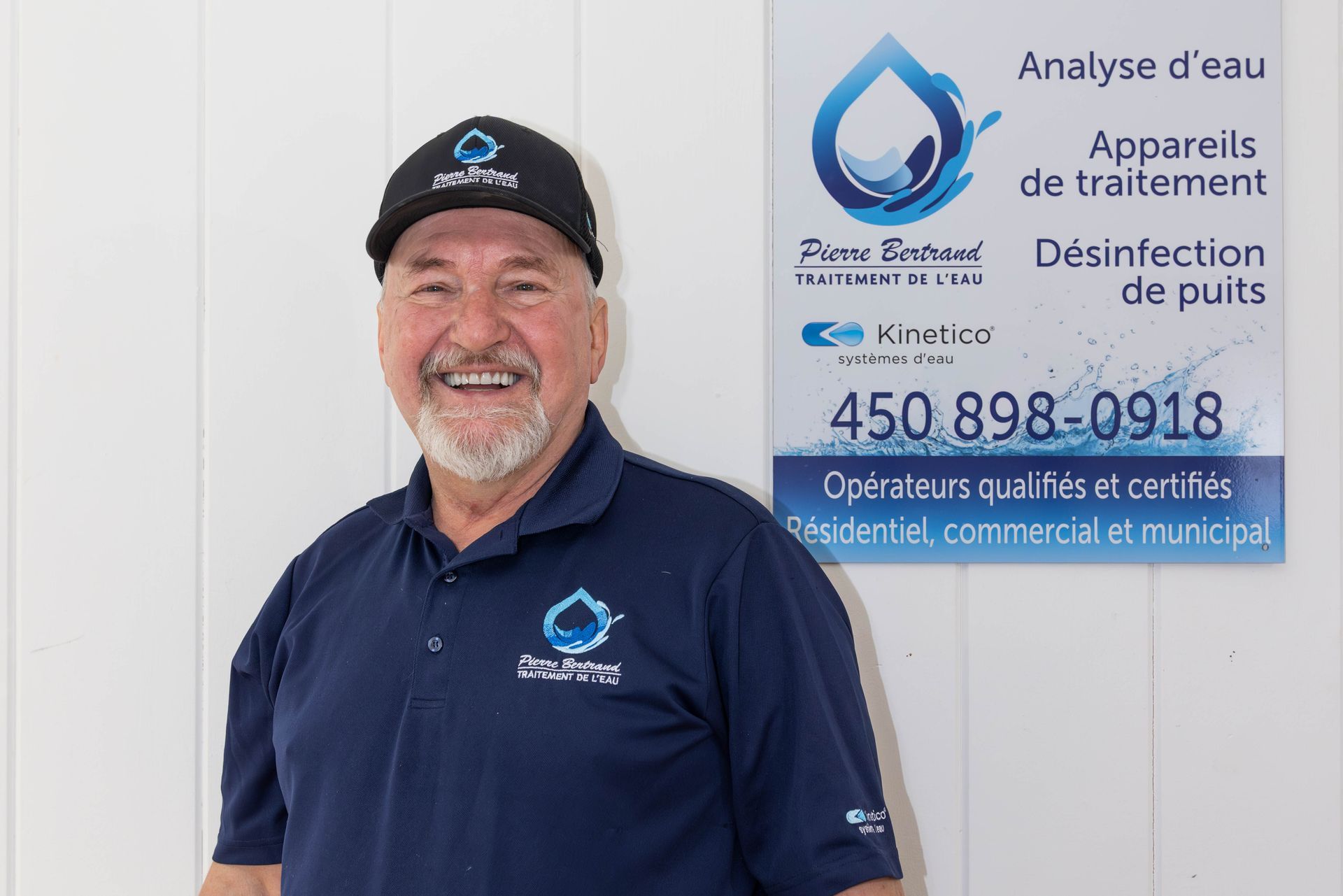 Un homme en chemise bleue et casquette sourit à côté d'un panneau concernant les services d'eau.