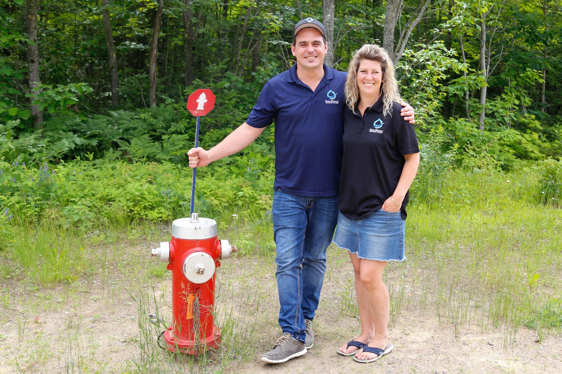 Un couple se tient à côté d'une borne d'incendie rouge et d'un panneau de signalisation de sentier pédestre, entouré de verdure.