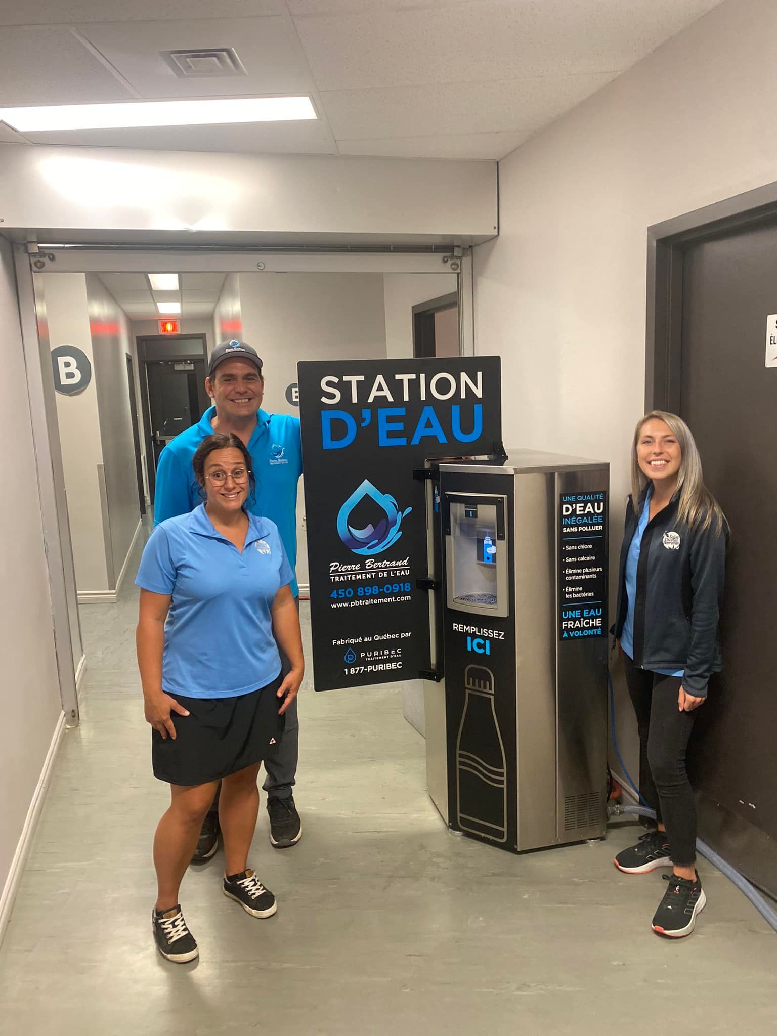 Trois personnes se tiennent près d'une fontaine à eau dans un couloir. La fontaine porte l'inscription « Station d'eau ».