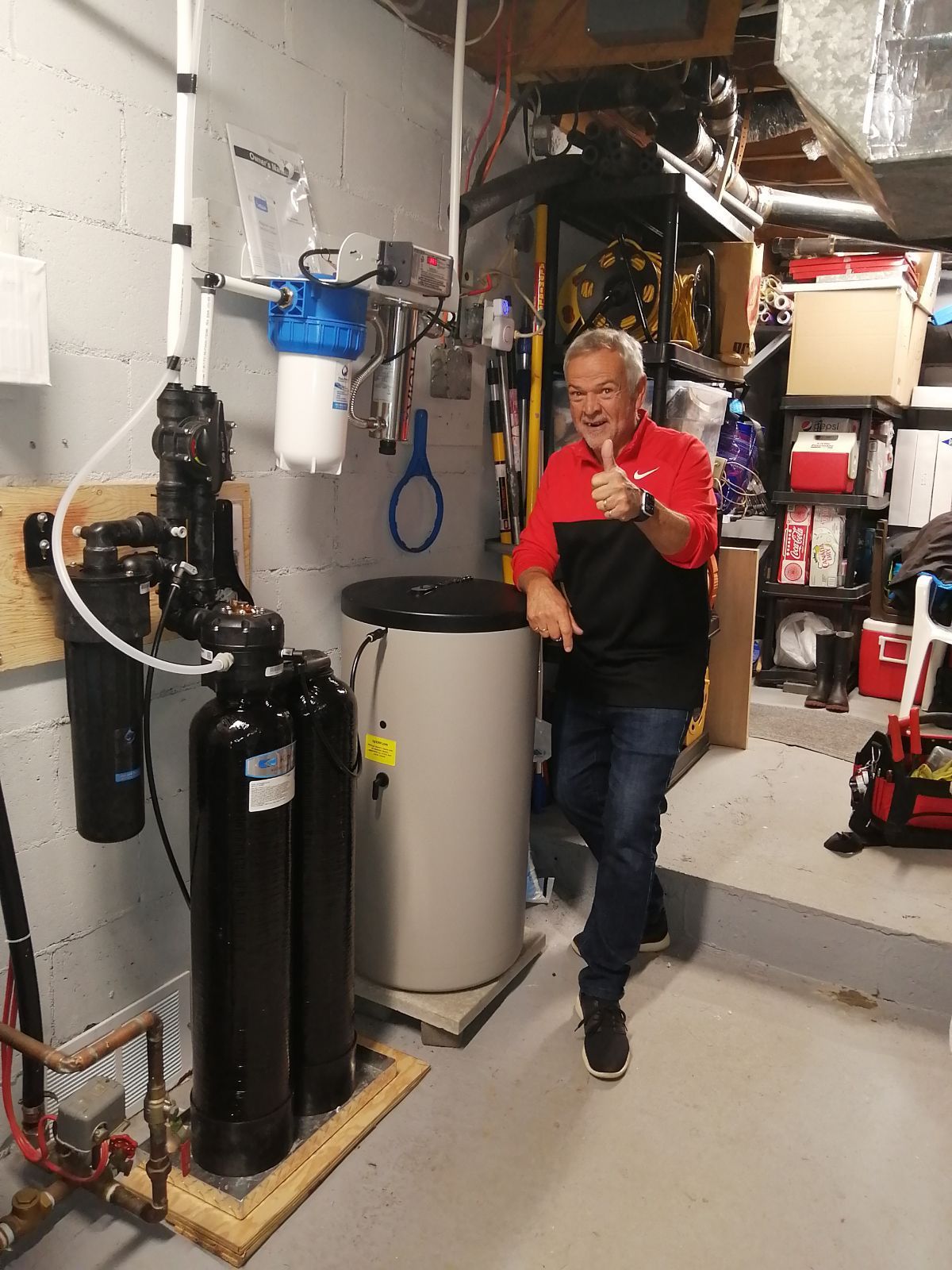 Un homme lève le pouce à côté d'un système de filtration d'eau dans un sous-sol.