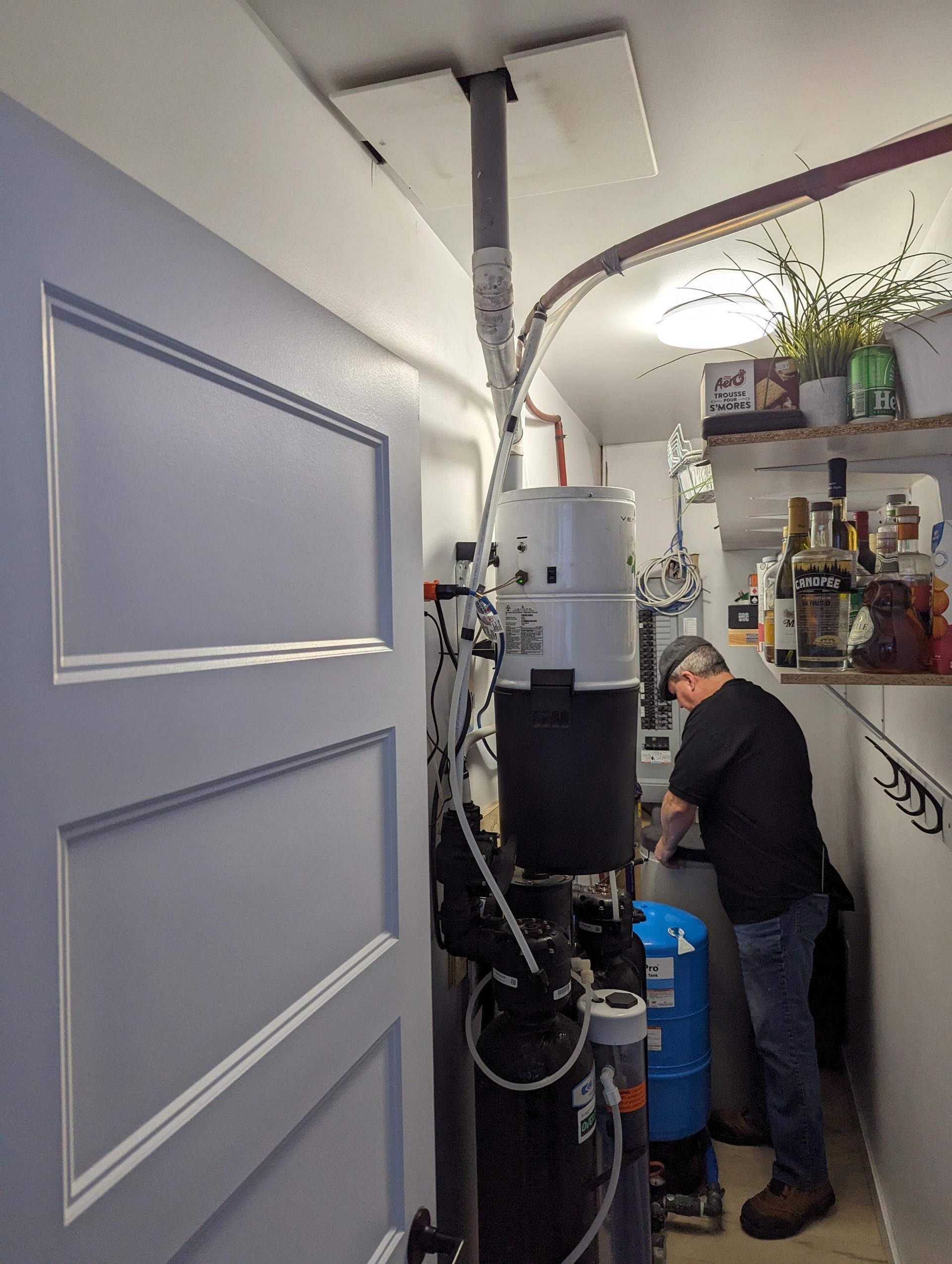 Un homme travaille sur un système de filtration d'eau près d'une porte. Réservoirs et tuyaux gris, noirs et blancs. Environnement : garage.