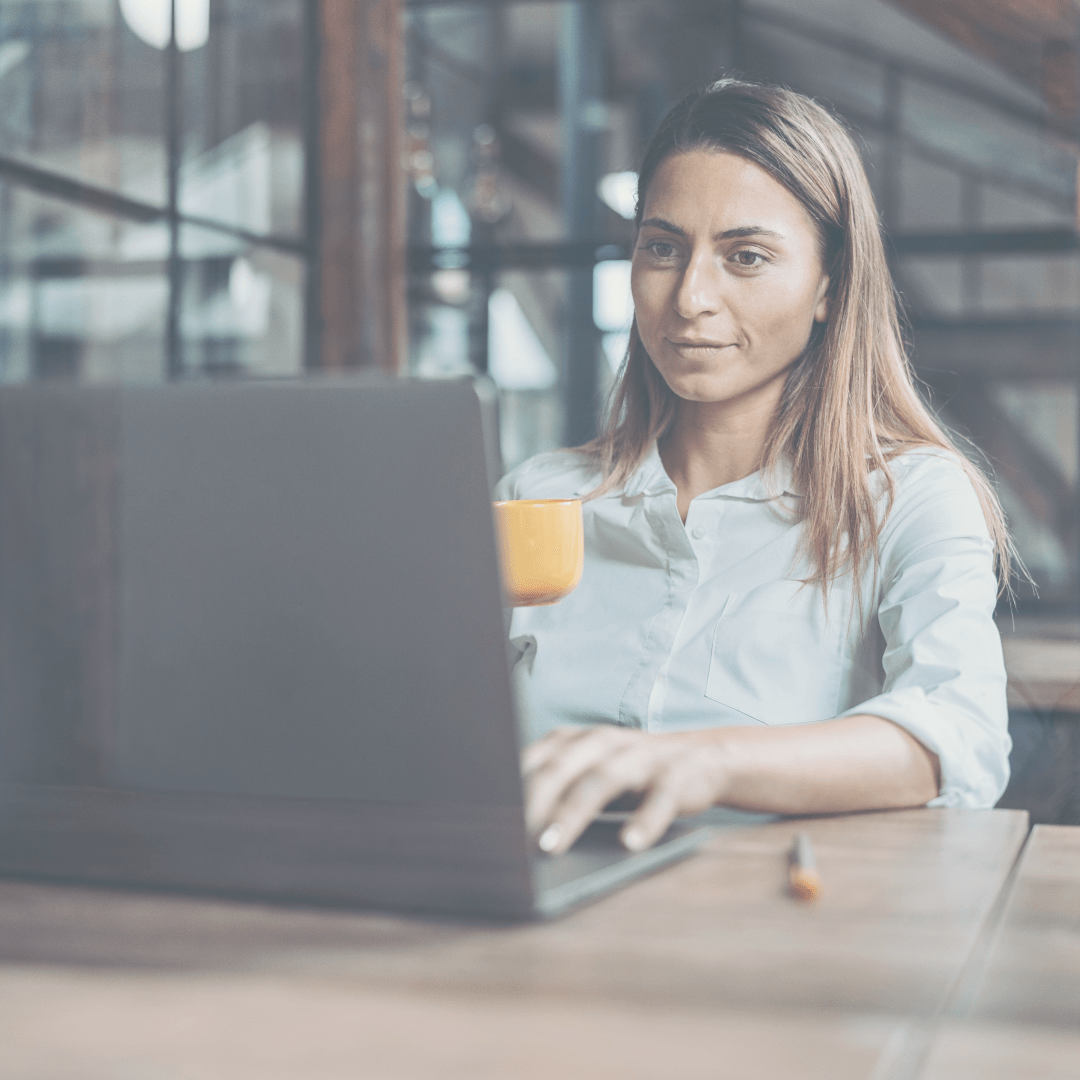 Vrouw in witte blouse, met laptop en koffiemok in haar hand, aan een houten tafel.