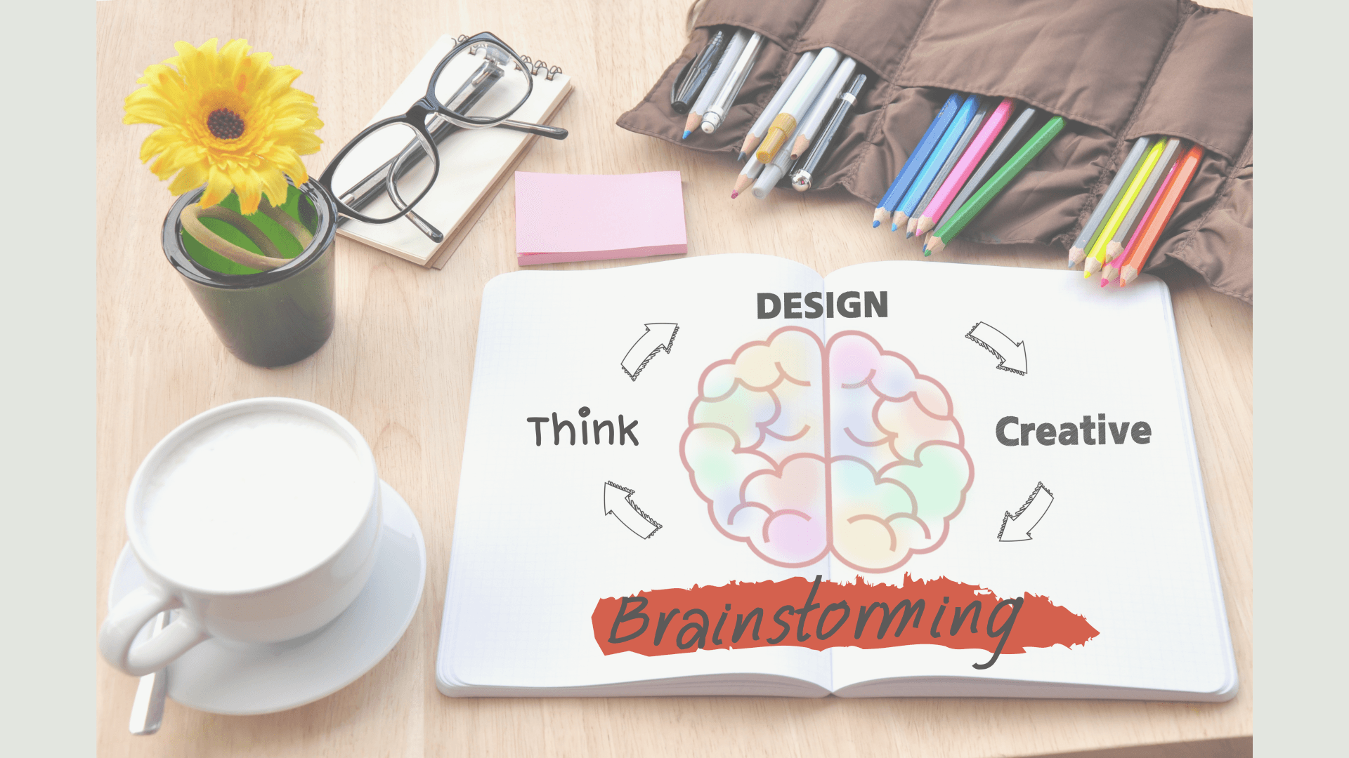 Opstelling voor een brainstormsessie met een afbeelding van een brein in een notitieboekje, pennen, een bril, koffie en een bloem.
