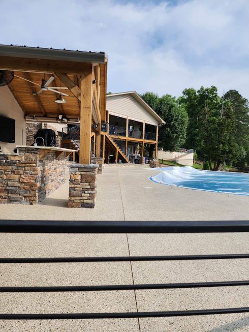 Stone patio with a wooden pavilion beside a blue swimming pool, trees in the background.