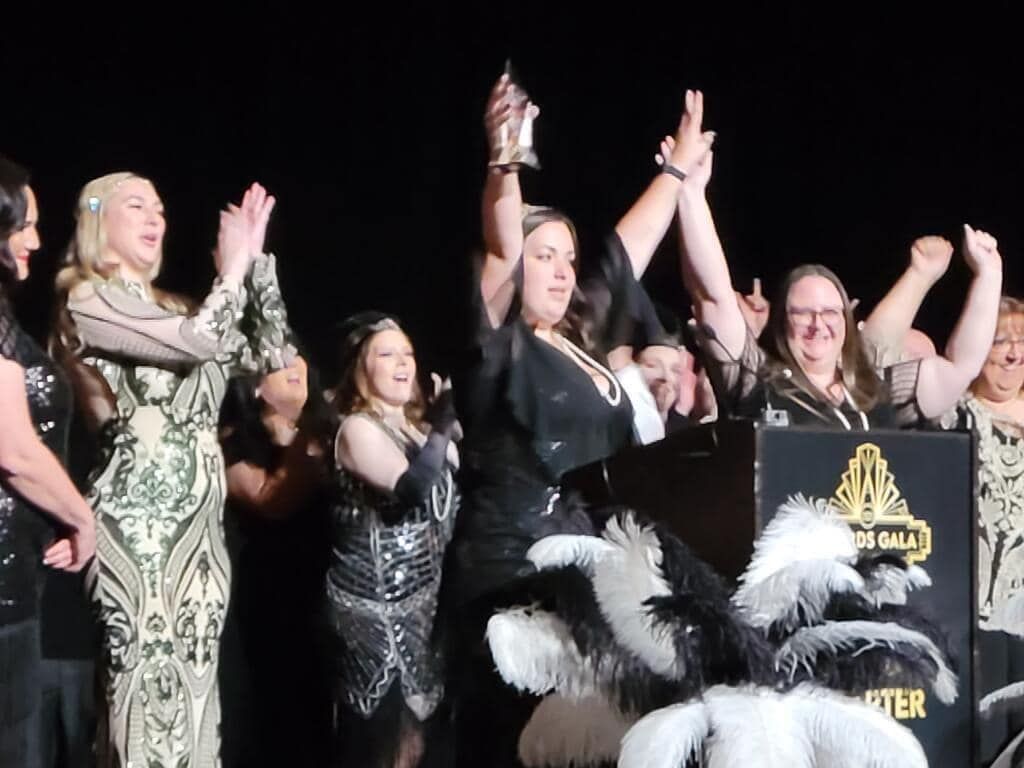 People cheering onstage beside a podium, one holding a trophy, under bright lights.