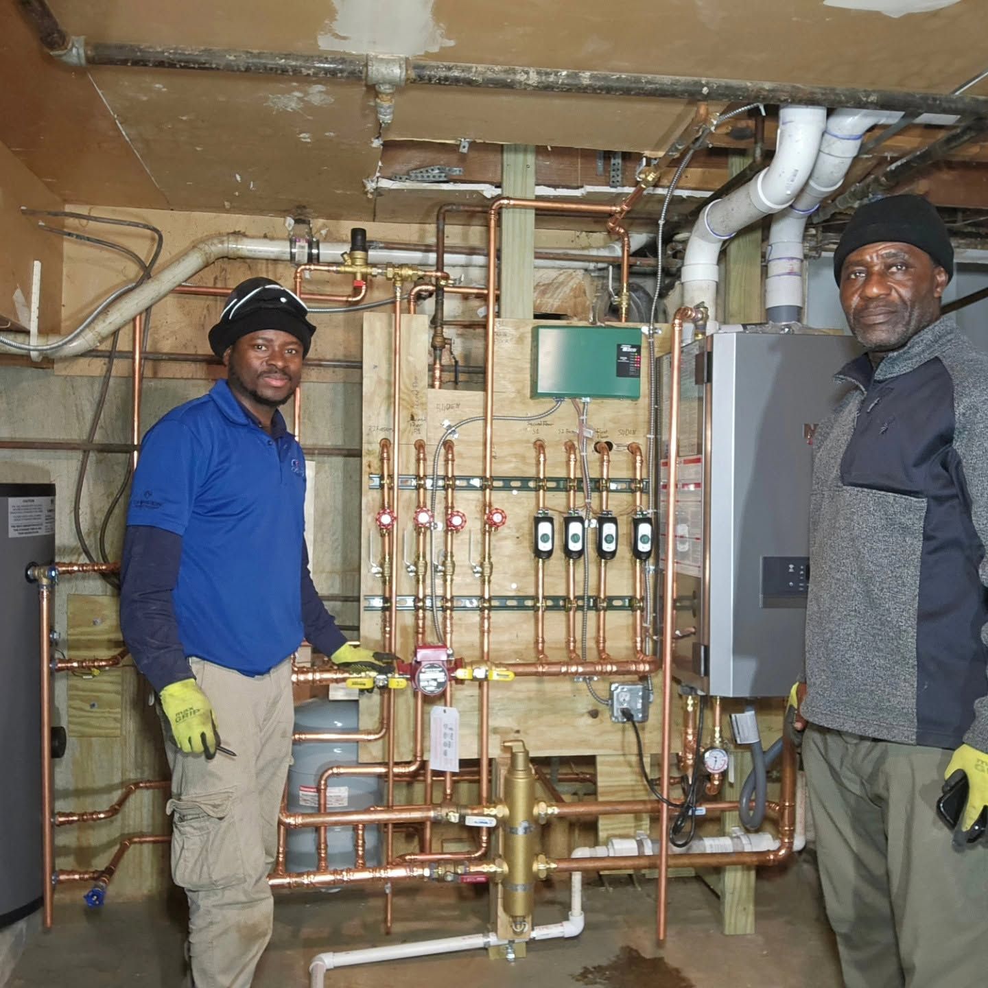 Two workers standing in a mechanical room beside plumbing and HVAC equipment