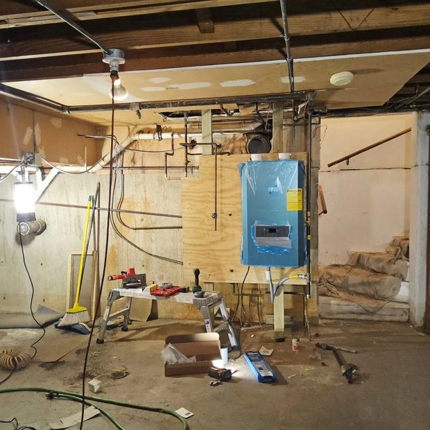 Basement under renovation with exposed ceiling, hanging wires, blue utility panel, and construction tools.