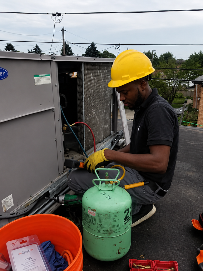 Worker in a yellow hard hat handling equipment beside a green gas cylinder outdoors.