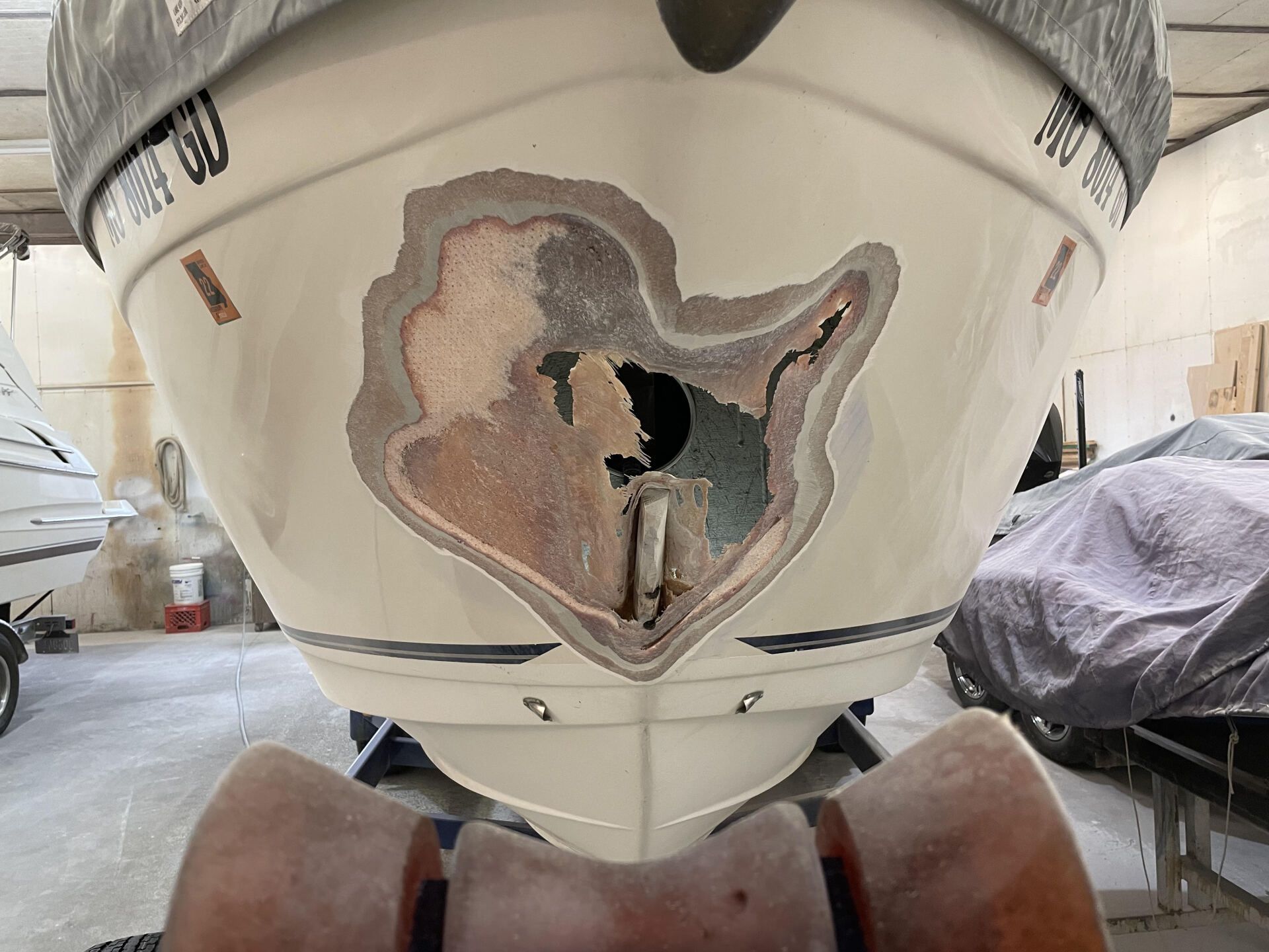 Boat hull with a large damaged, rusted patch in a boatyard, viewed from the front.