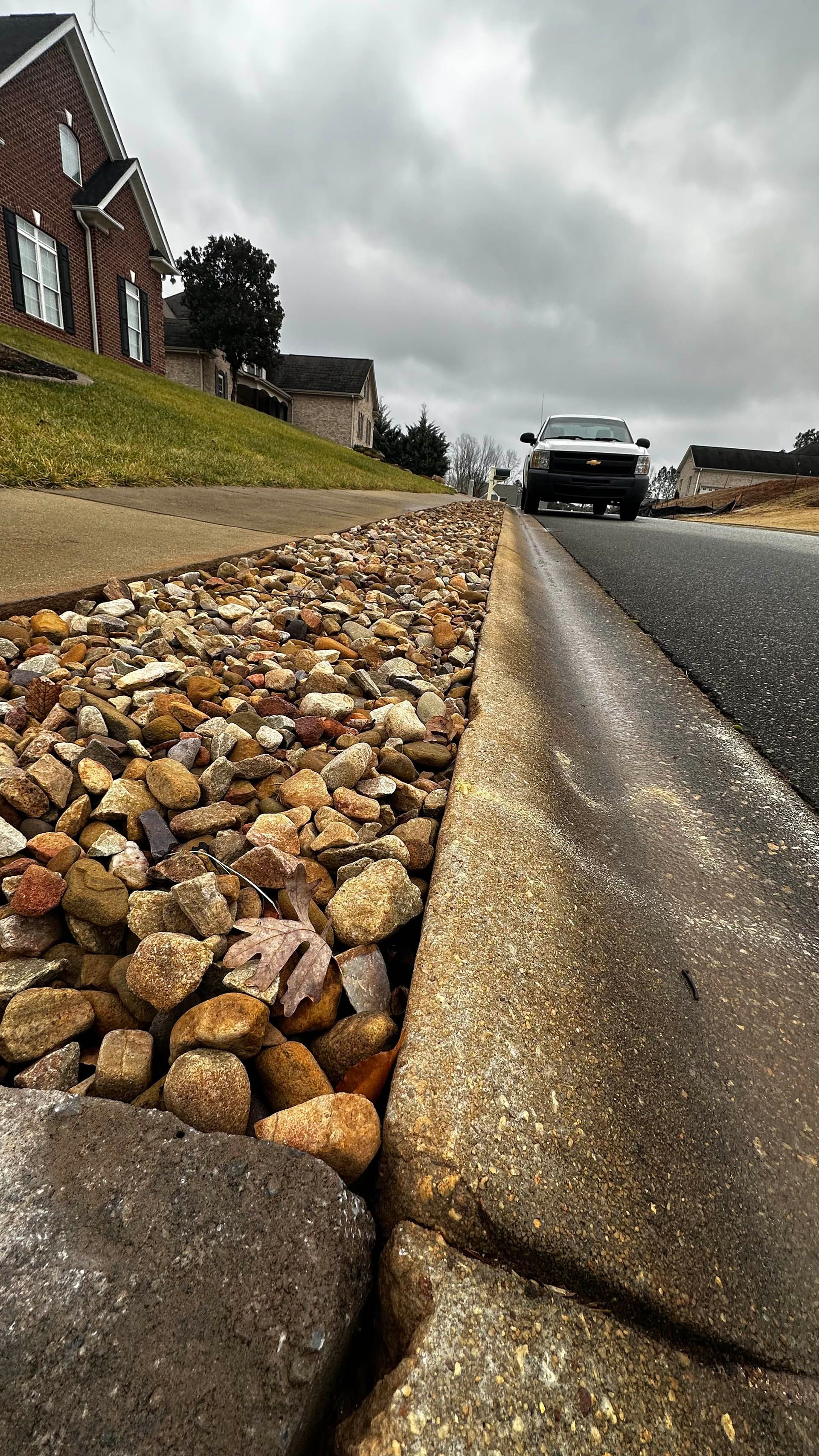 Low-angle view of a rocky roadside gutter beside houses, with a car approaching on a cloudy street.