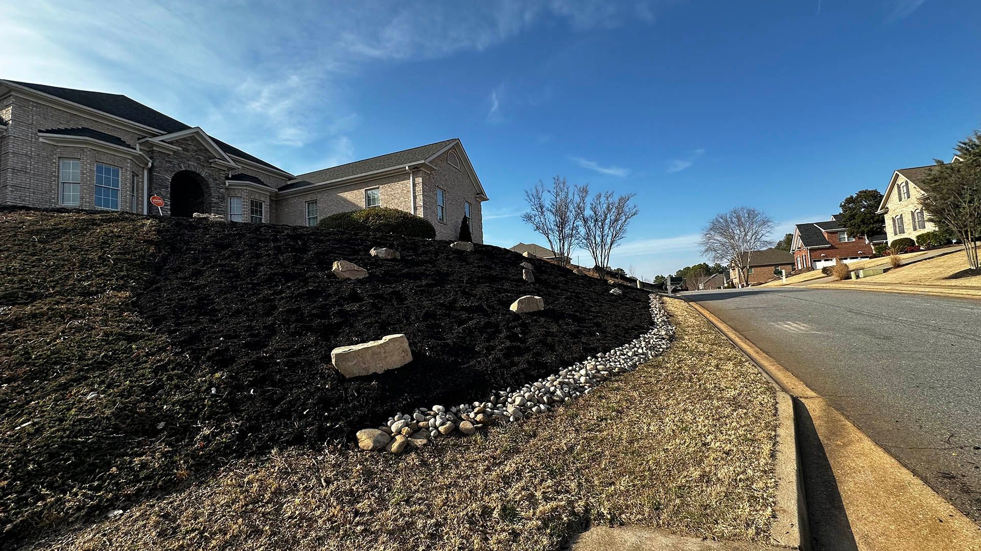 Sunlit suburban street beside a stone retaining wall and houses under a clear blue sky
