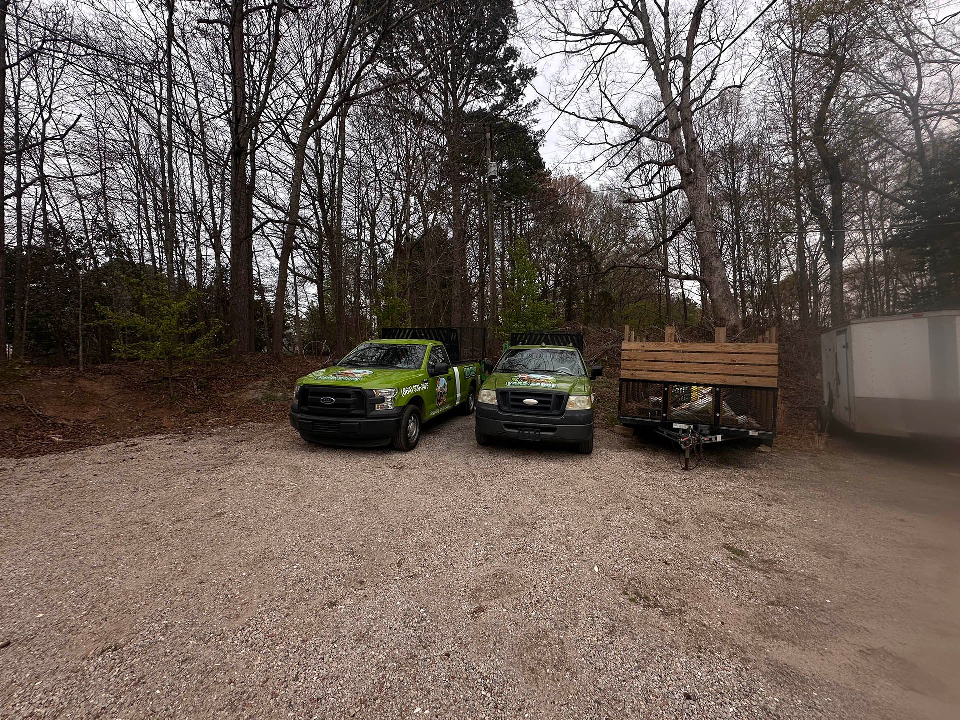 Three vehicles parked on a gravel lot beside leafless trees, including two green trucks and a wooden trailer.