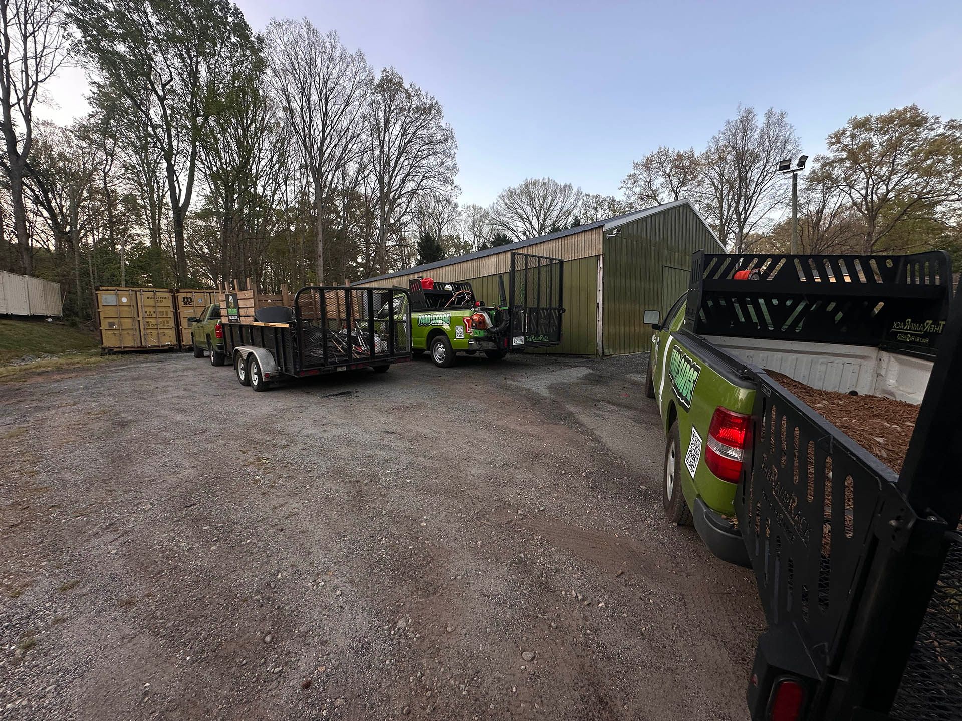Workers and utility trucks in a muddy yard beside stacked materials and trees