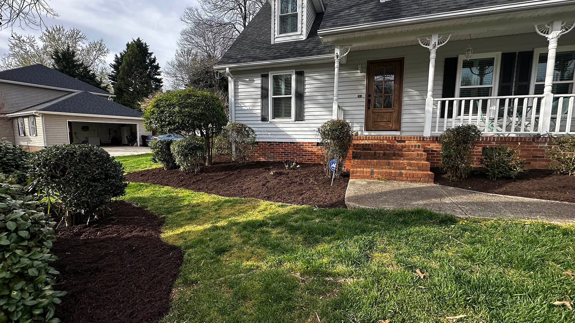 Front yard of a white house with a porch, brick steps, trimmed shrubs, and green lawn in sunlight