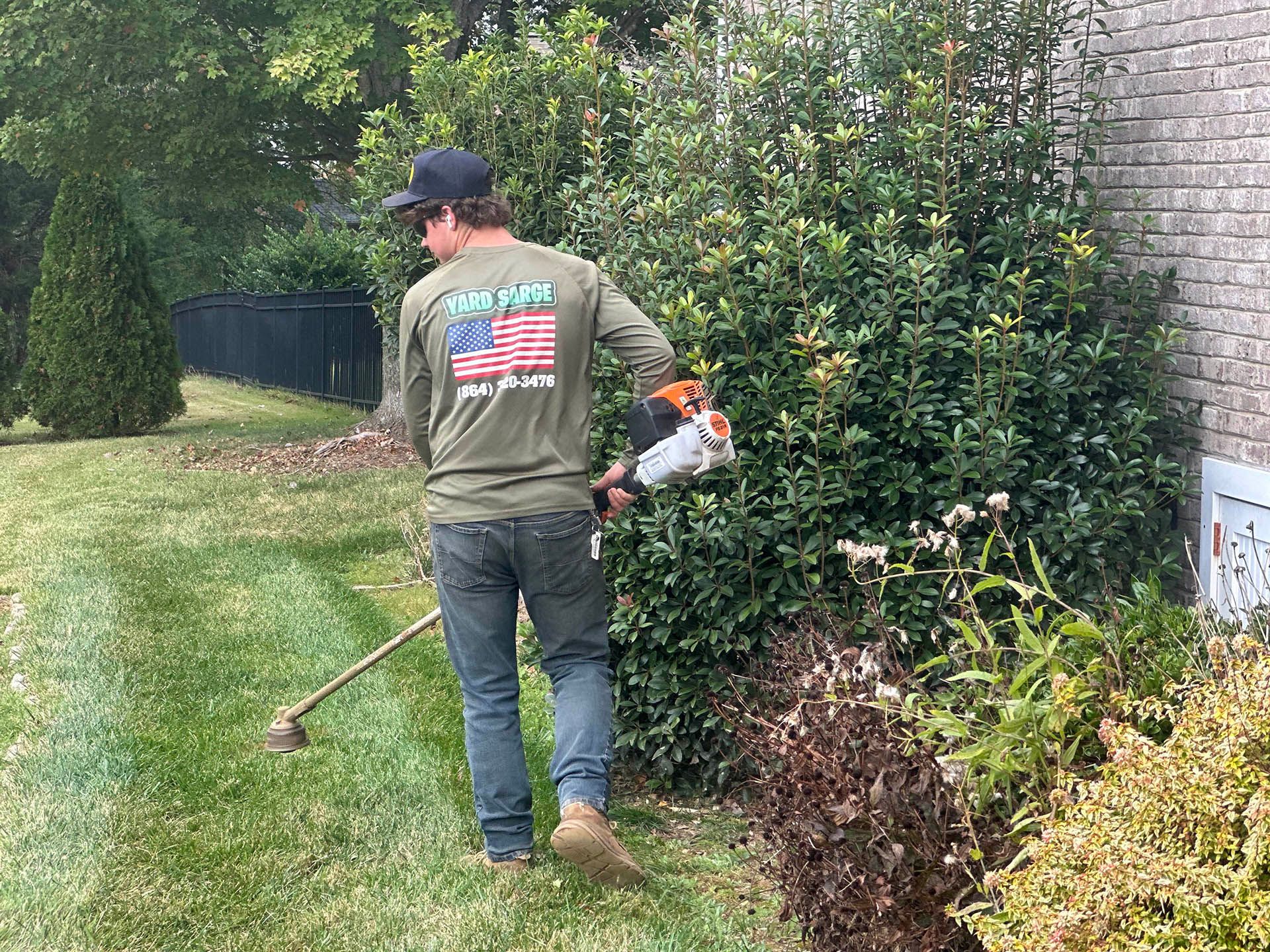 Man trimming hedges along a lawn with a string trimmer near a fence and bushes.