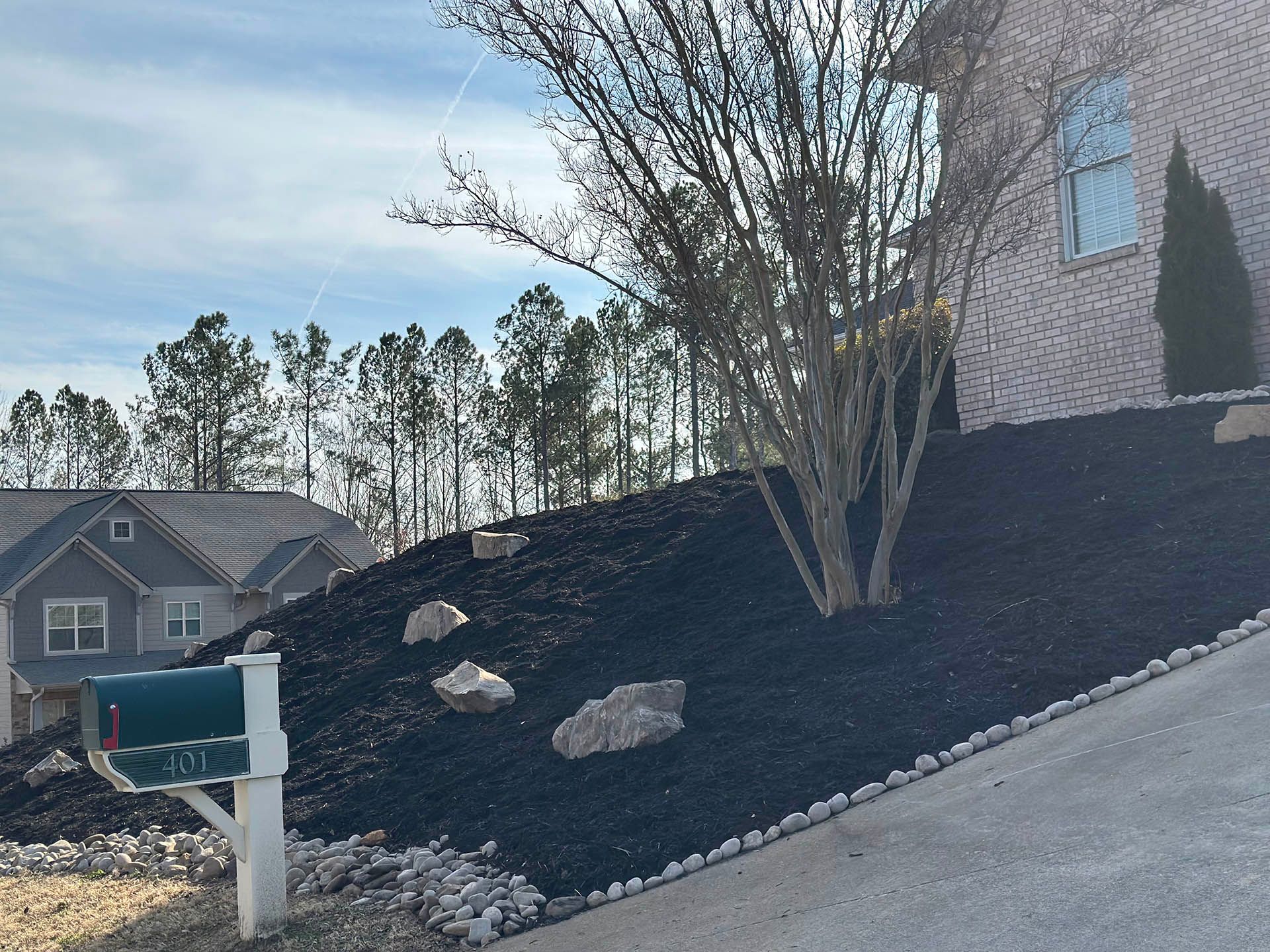 Suburban house with steep driveway, dark mulch landscaping, mailbox, rocks, and bare trees under a blue sky