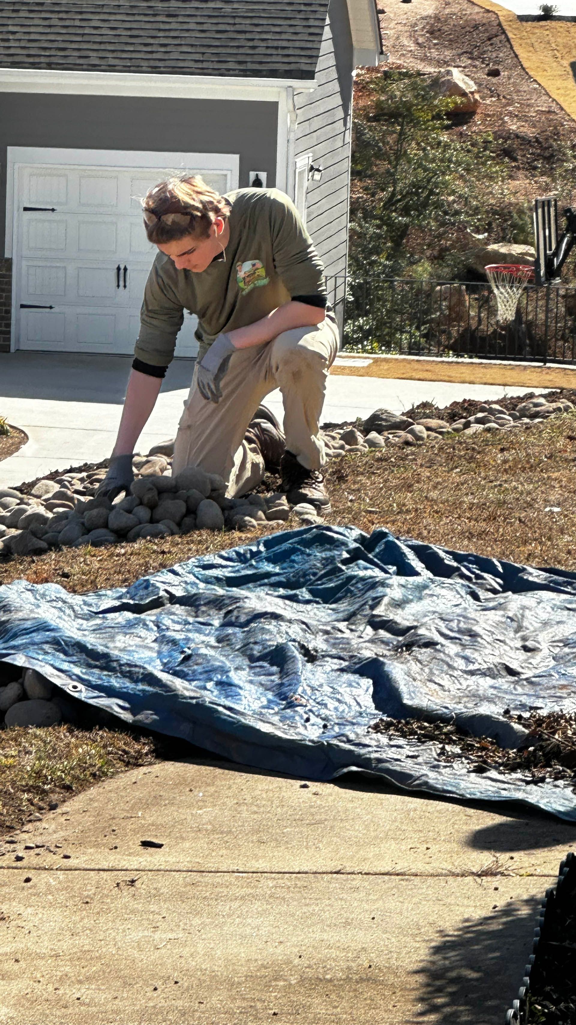 Person kneeling on driveway spreading rocks over landscaping fabric beside a house in daylight