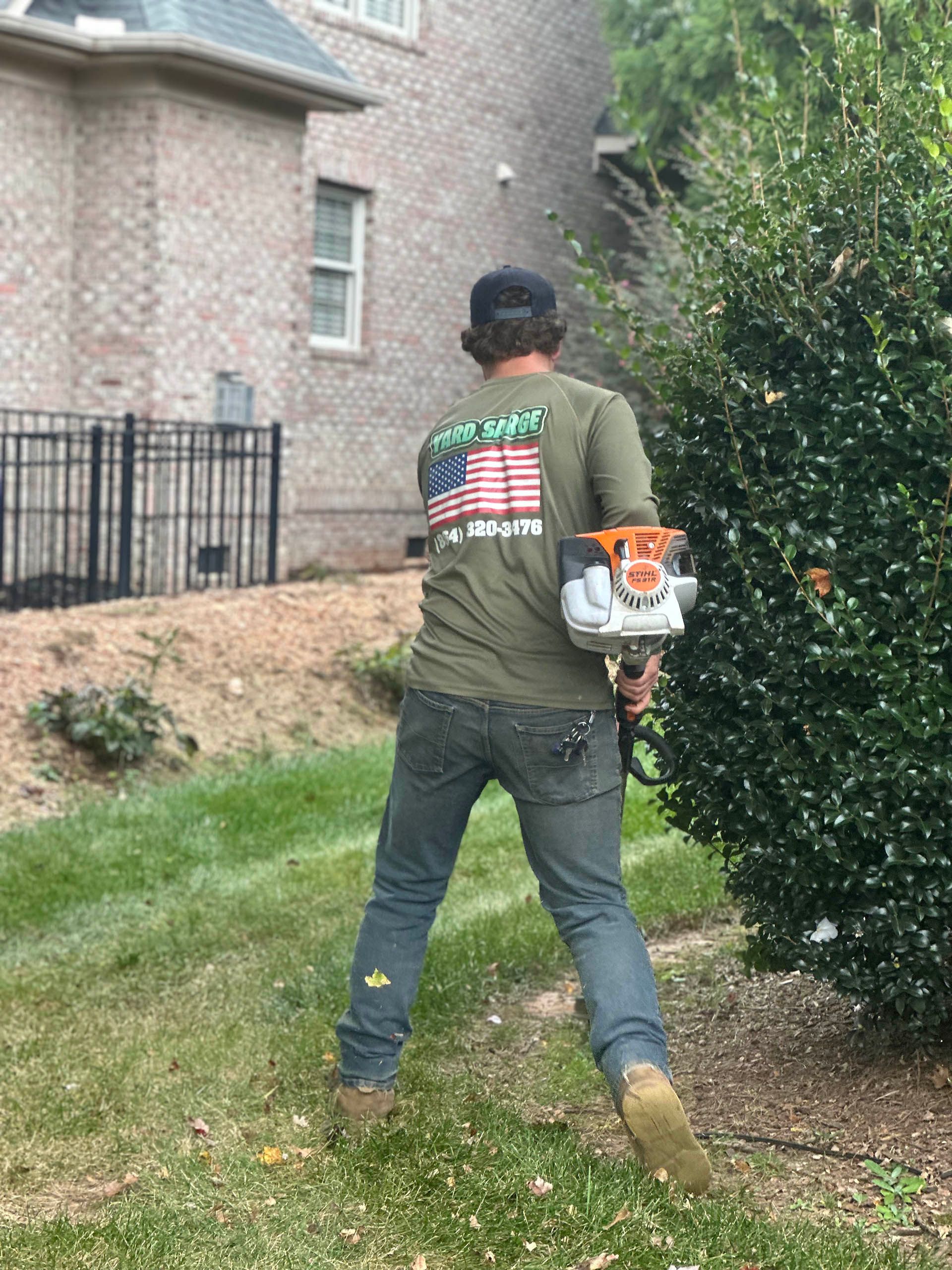 Person using a leaf blower in a residential yard beside a brick house and shrubbery