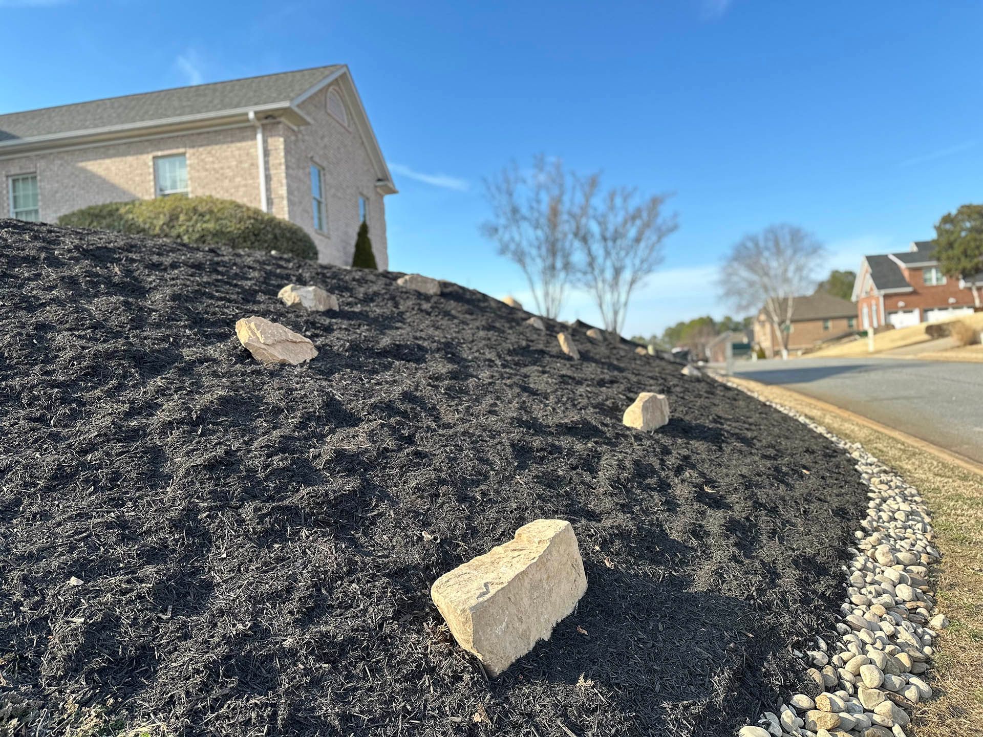 Landscaped roadside mound of dark mulch with rocks beside a suburban street and house