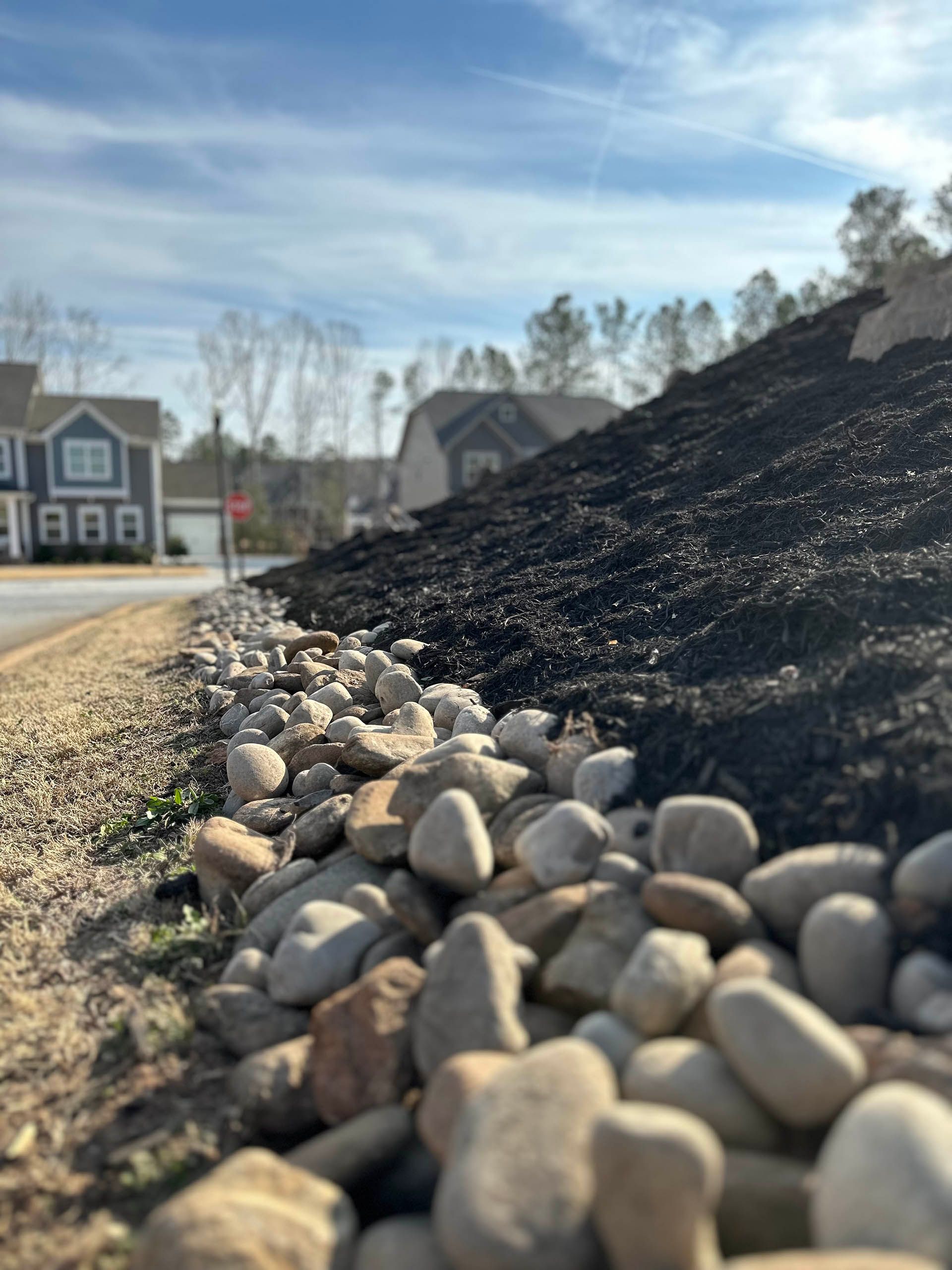 Rocks and dark mulch lining a suburban roadside with houses in the background under a blue sky