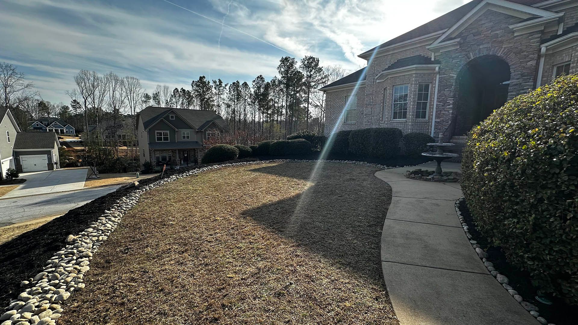 Stone house and curved walkway in a sunny yard with leaf-covered mulch and a lens flare in the foreground
