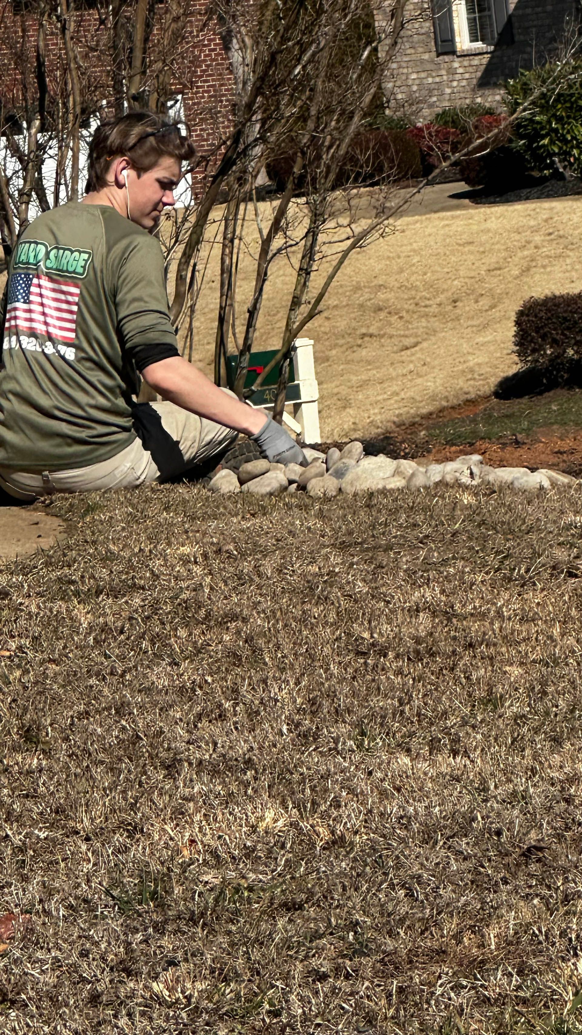 Person kneeling in a yard, working with a trowel near a small post and rocks on a sunny day