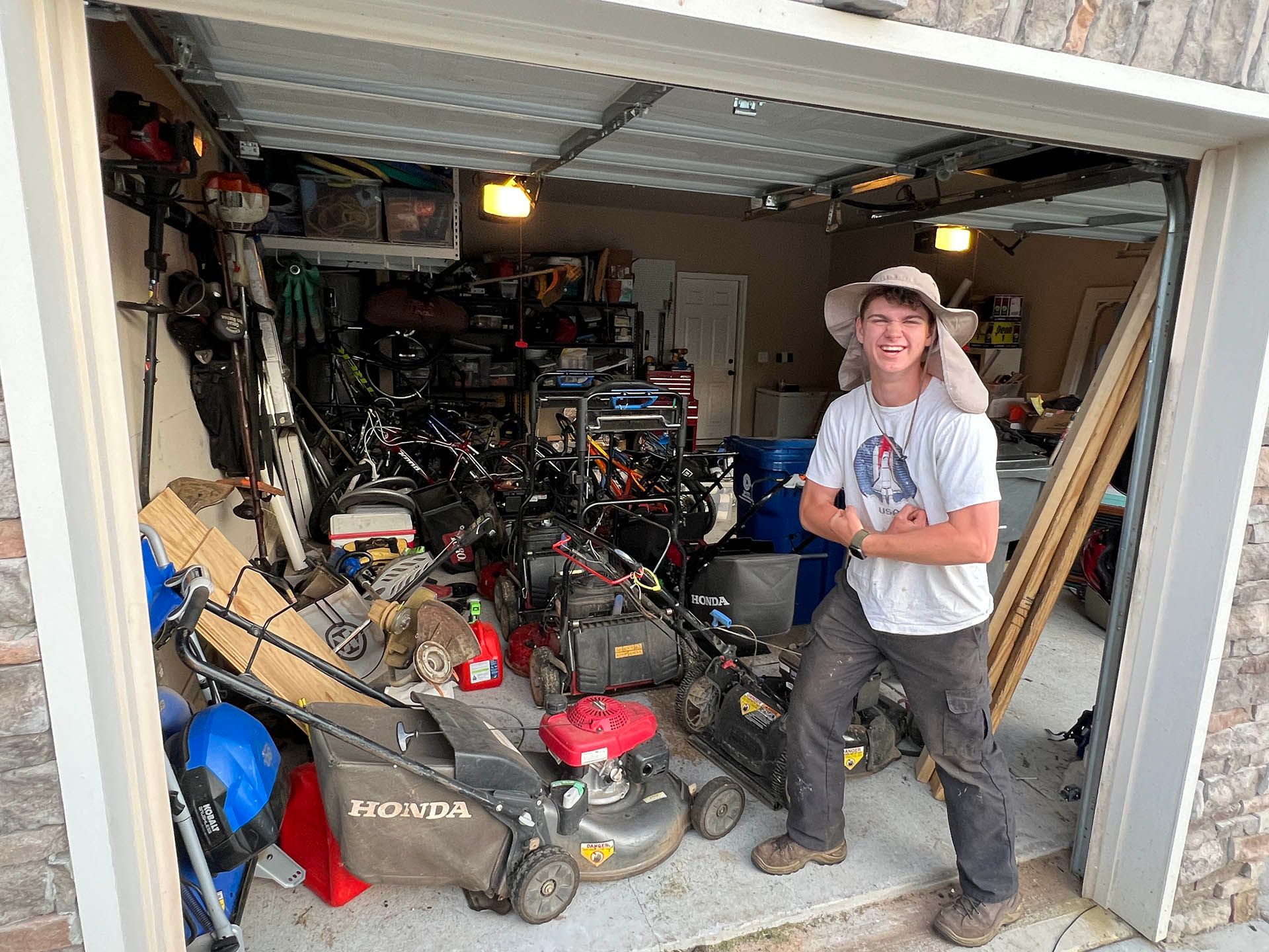 Person standing in a cluttered garage filled with lawn equipment and tools, holding a drink.
