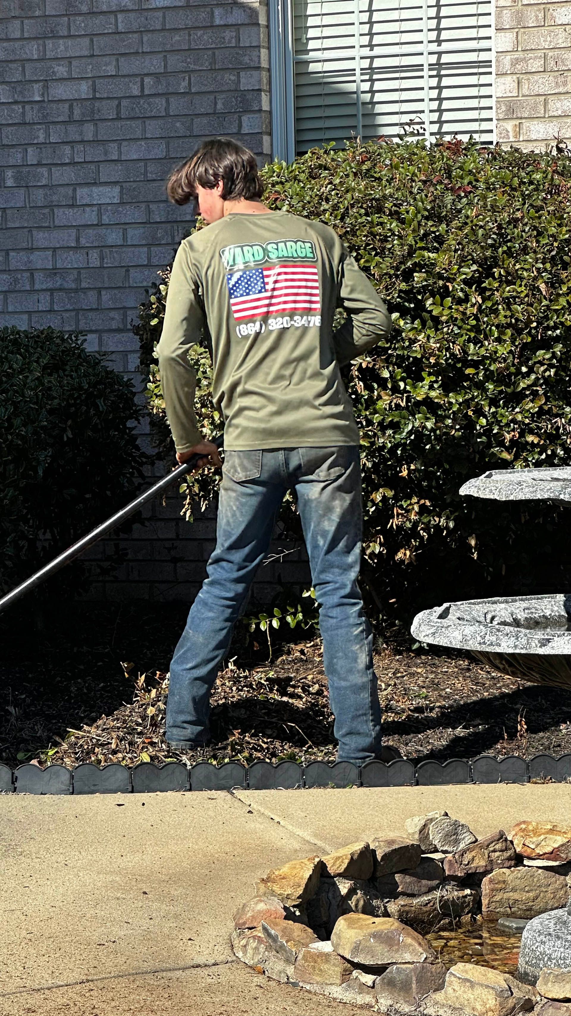Person watering shrubs in a garden, wearing a green shirt with an American flag on the back.