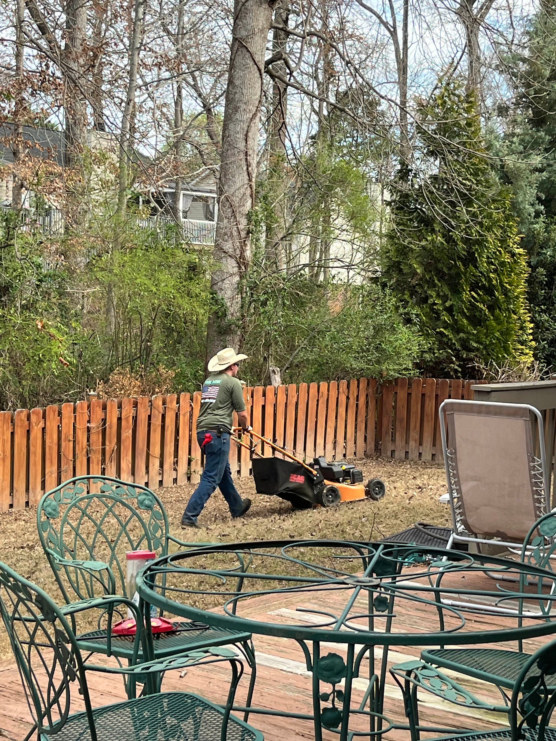 Person mowing a grassy yard beside a wooden fence, with patio chairs in the foreground.