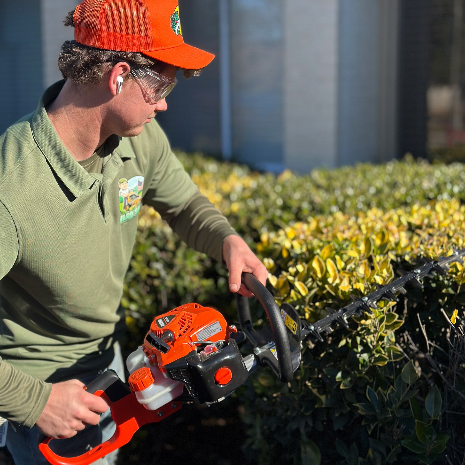 Worker trimming hedges with a red-orange hedge trimmer beside a landscaped bush