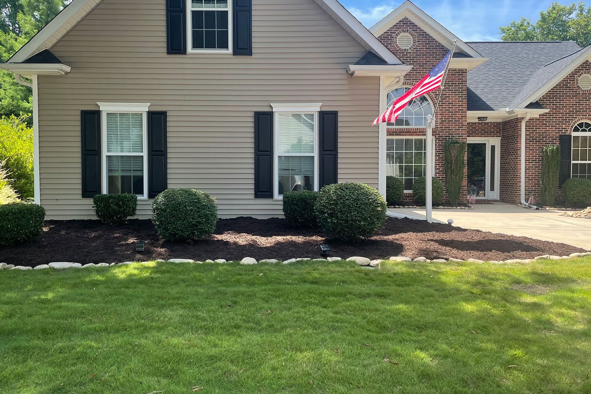 Suburban tan house with black shutters, American flag, and a green front lawn