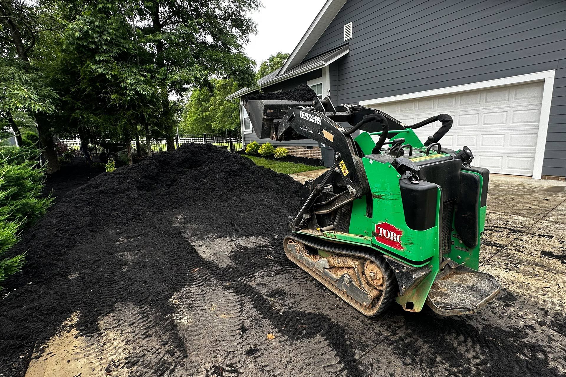 Green skid steer moving a large pile of black dirt beside a white garage