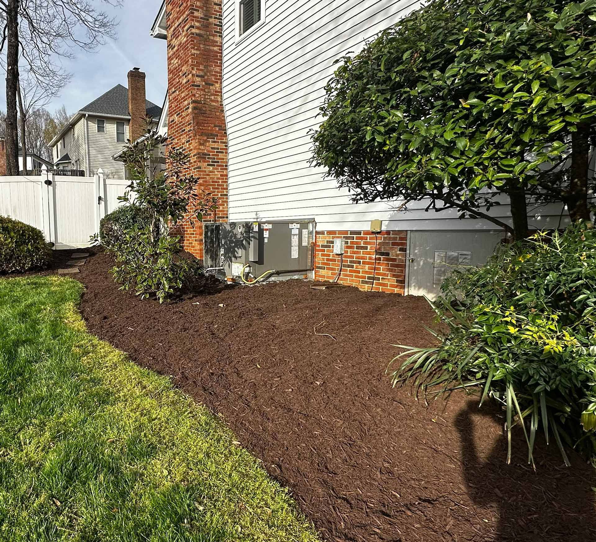 Fresh brown mulch bed along a house, beside a green lawn and shrubs, with a tree and white fence nearby