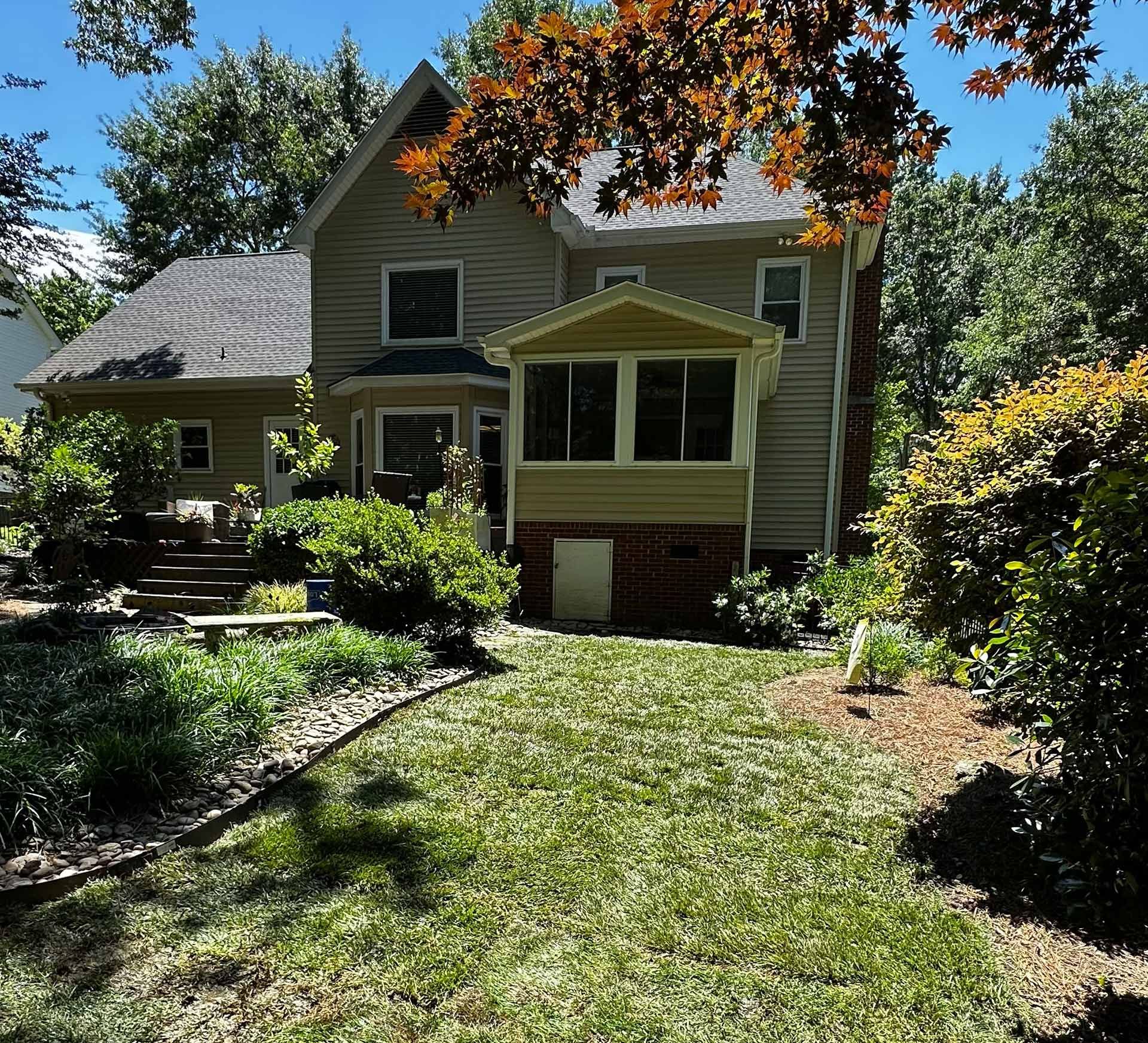 Sunlit house with a green lawn, shrubs, and a tree canopy in the foreground