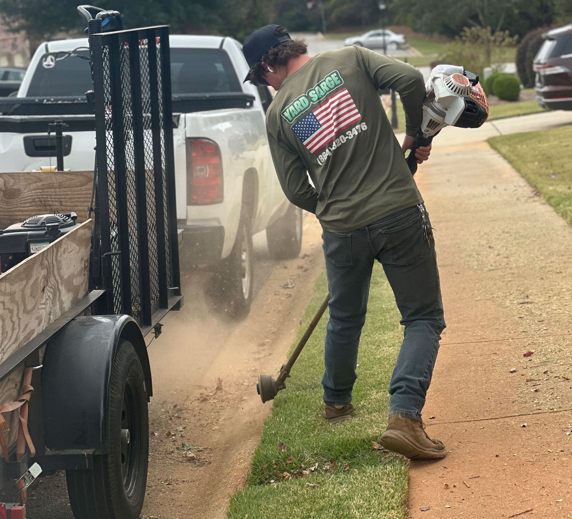 Worker using a gas trimmer to edge grass beside a sidewalk near a white pickup truck and trailer.