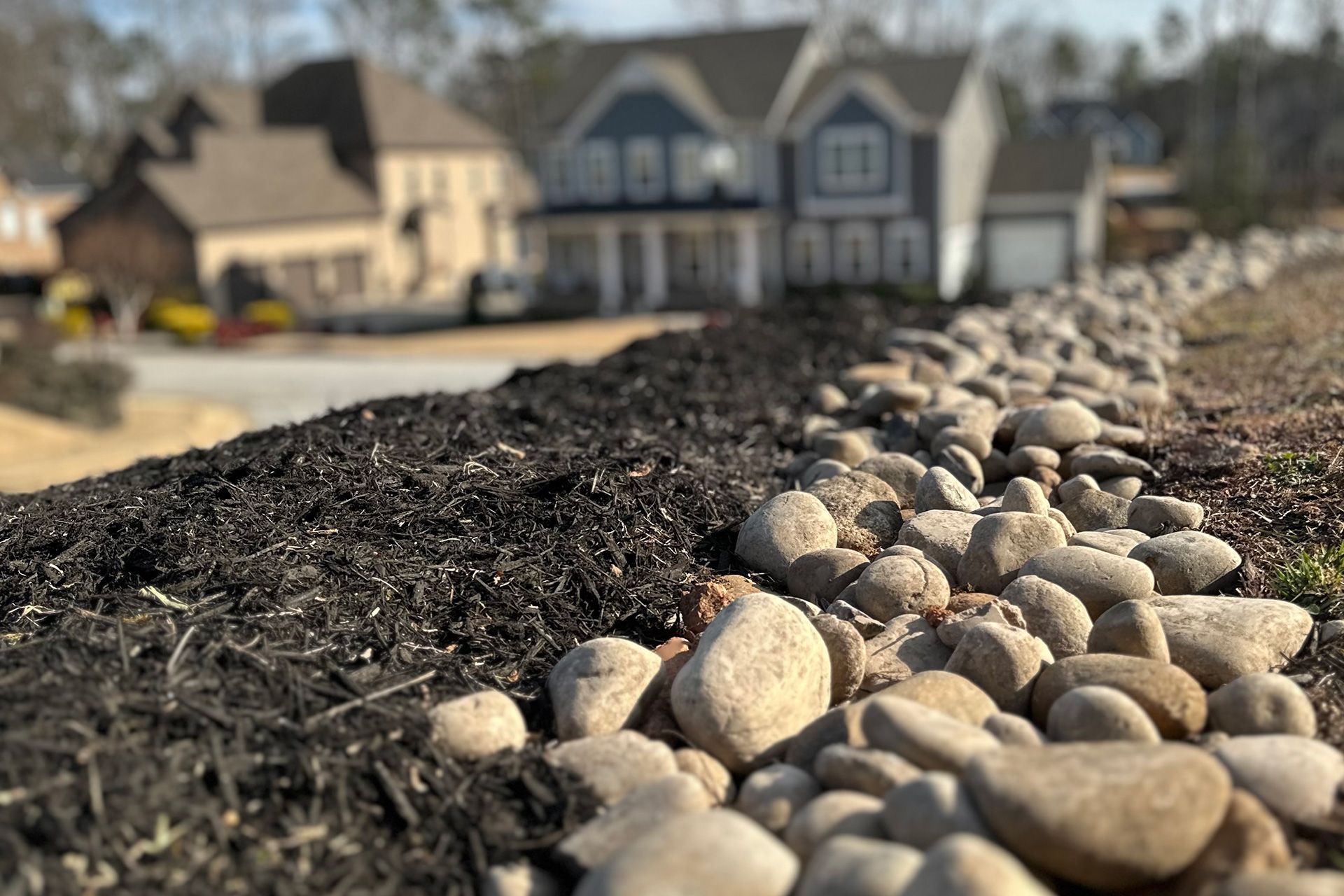 Mulch and river rocks lining a landscaped front yard with houses blurred in the background