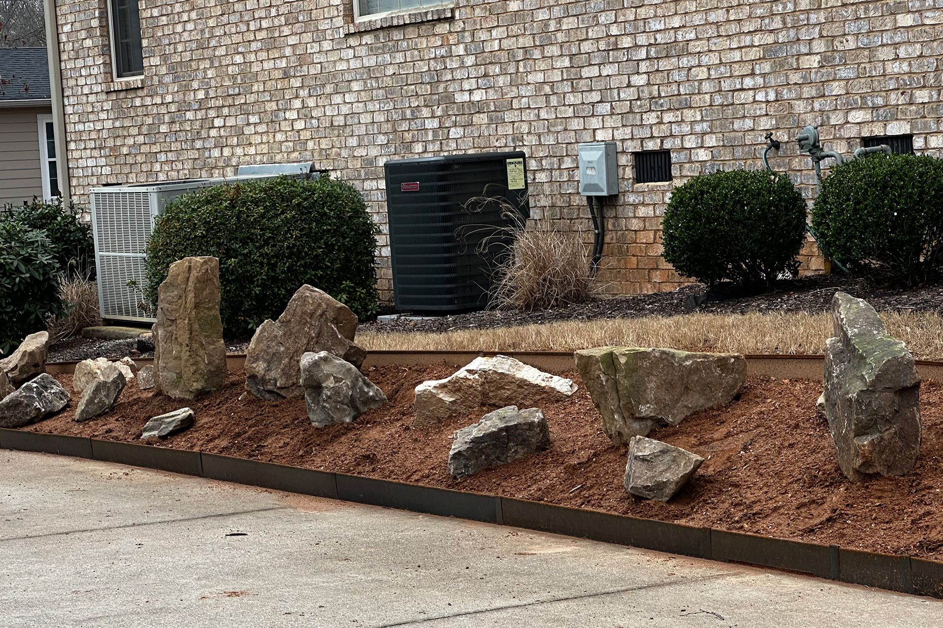 Landscape bed with large rocks, mulch, shrubs, and utility boxes against a stone building exterior